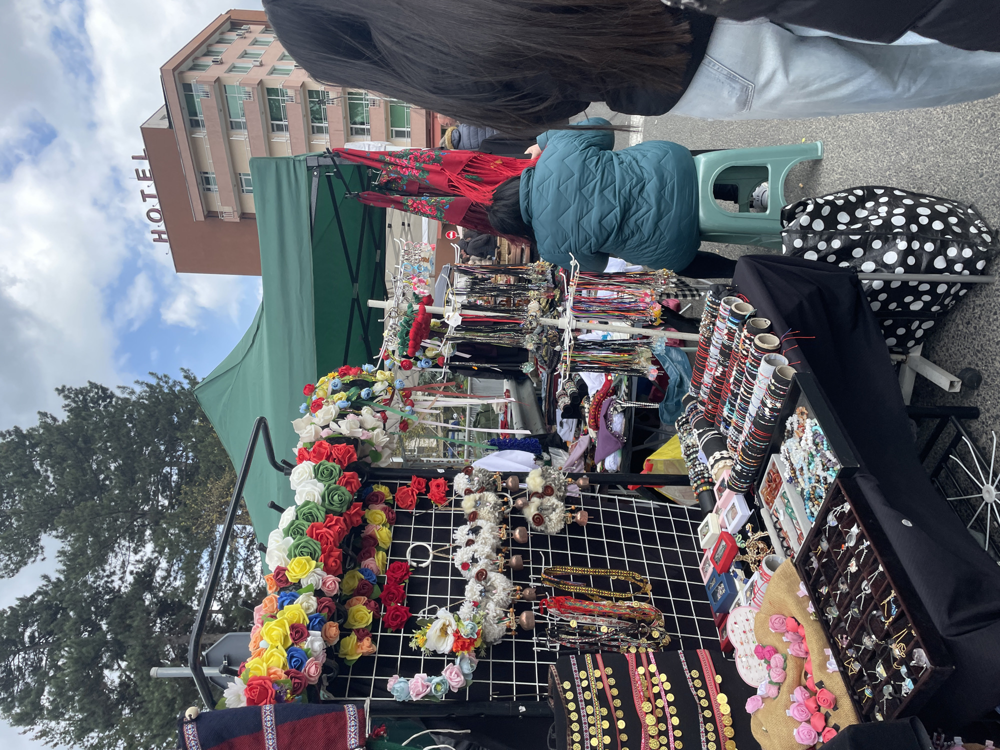 The image captures an outdoor market stall in Zlatograd, Bulgaria, displaying various jewelry and accessories. Colorful flower crowns and fabric headbands are arranged on a wire grid. Bracelets, earrings, and necklaces are displayed on a table covered with black fabric. A customer in a blue jacket browses the merchandise while seated on a green plastic stool. Another person with long dark hair stands nearby, possibly a companion or another customer. In the background, a hotel building with the word "HOTEL" vertically aligned on its side is visible against a partly cloudy sky. The scene conveys a casual atmosphere of a local market.