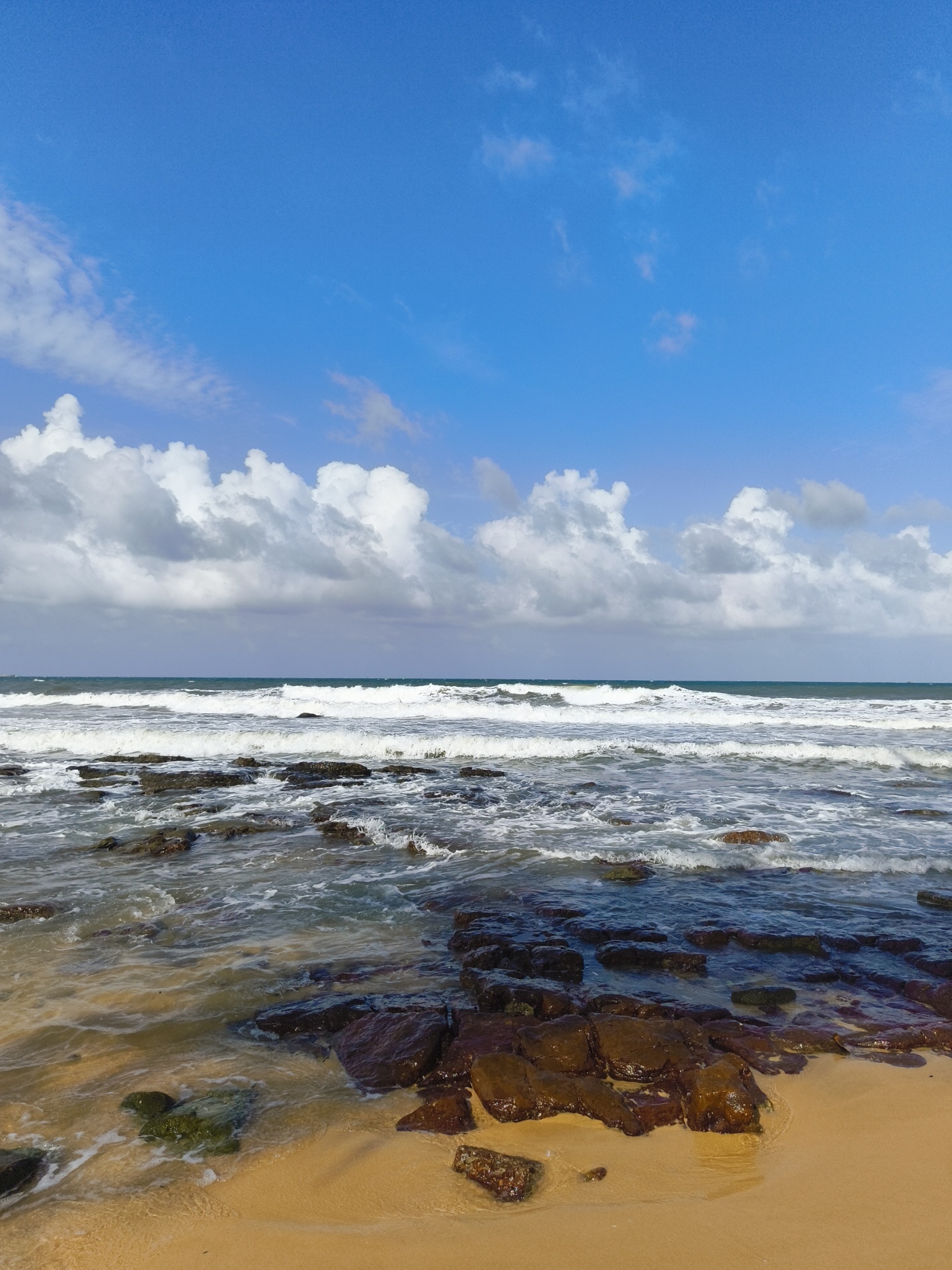 This is a landscape photograph of a rocky beach on Phu Quoc Island, Vietnam. The image captures a portion of the ocean with waves breaking over dark, wet rocks that are partially submerged in the water and interspersed with sand. The beach itself is composed of golden sand, with more rocks and shallow water in the foreground. The sky above is a vibrant blue, dotted with fluffy white clouds. The overall scene depicts a natural coastal environment.