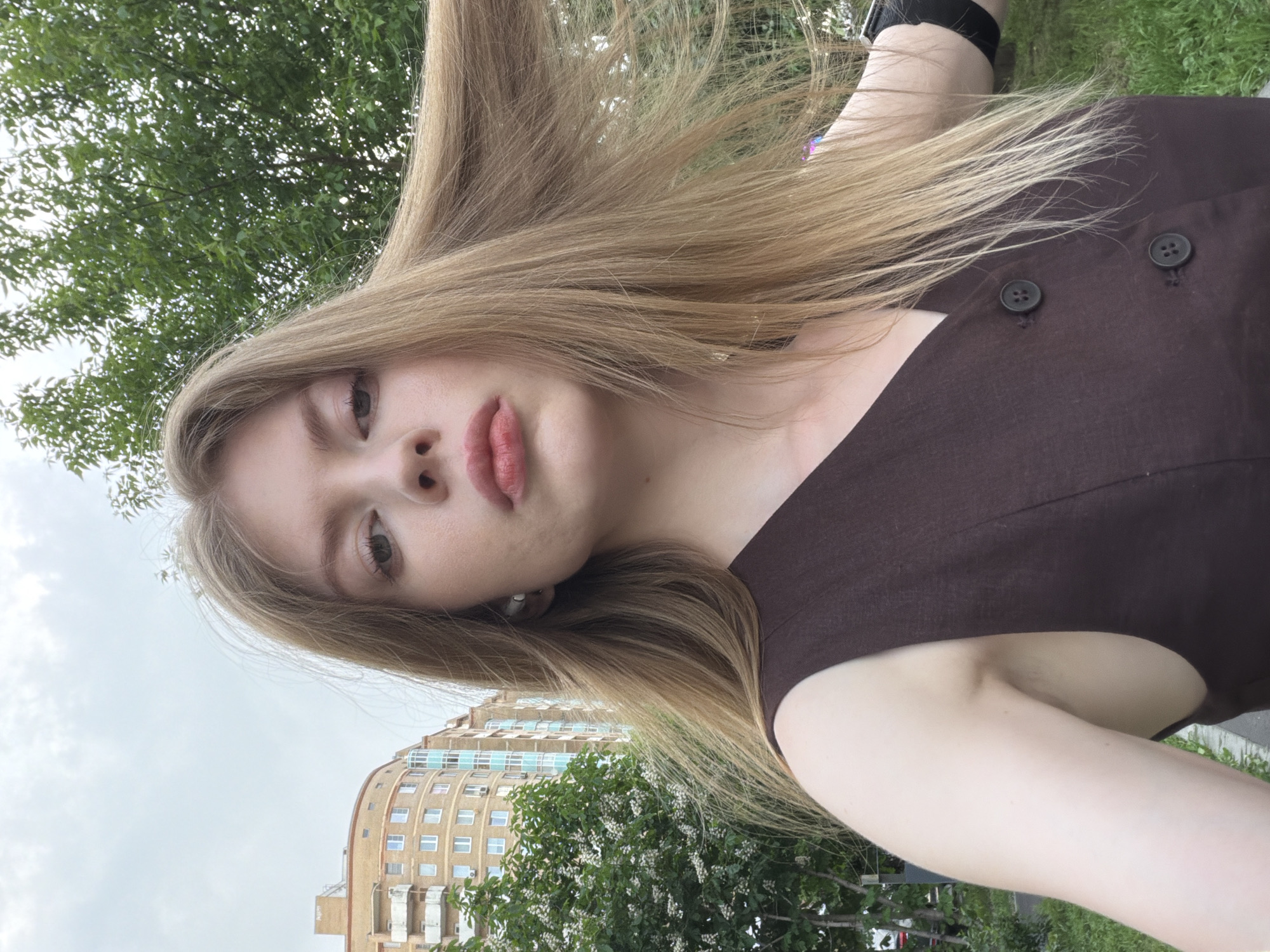 The image features a young woman with long, light-brown hair, posing in a selfie-style shot outdoors. She is wearing a dark-brown top with two buttons visible on the shoulder. The background includes a residential building with multiple windows, suggesting an urban environment. Green trees and foliage are also visible, indicating the presence of a park or green space nearby. The scene seems to be located in a park. The specific location is Moscow, Russia.