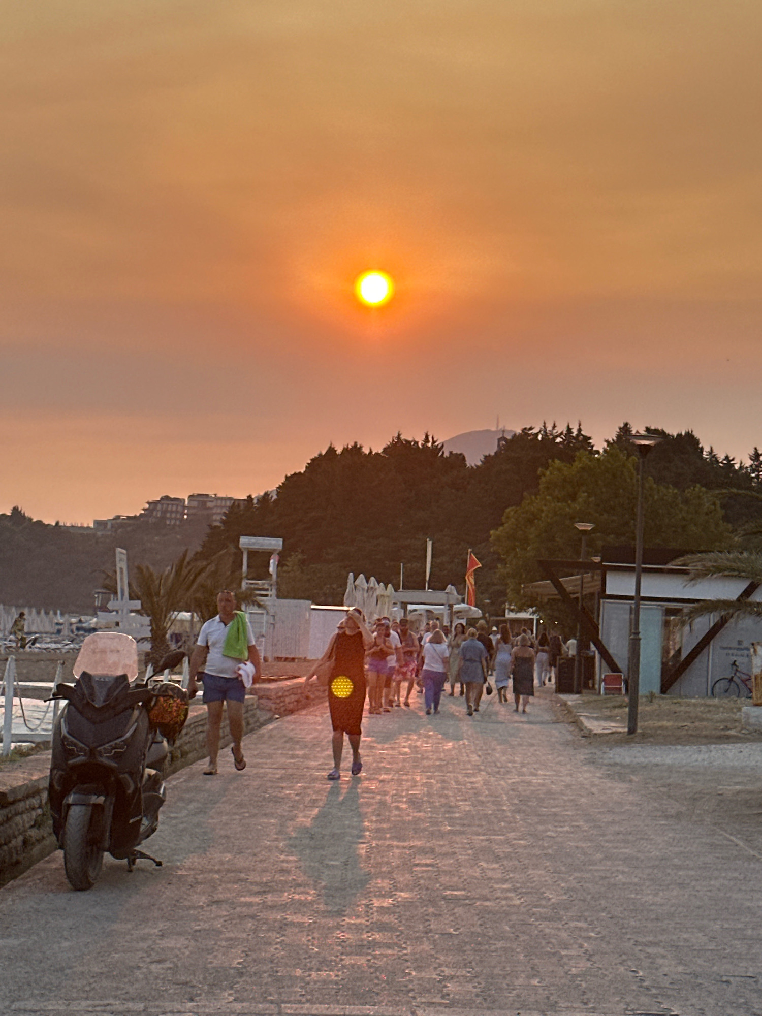 The image depicts a sunset scene on a promenade in Boreti, Montenegro. A group of people are walking along a cobblestone path. In the foreground on the left, a dark-colored scooter is parked. In the background, there are trees and buildings on a hillside, and the sun is setting, casting a warm orange glow over the scene. Beach umbrellas are visible on the left side of the promenade. The sky is hazy, contributing to the soft, diffused light.