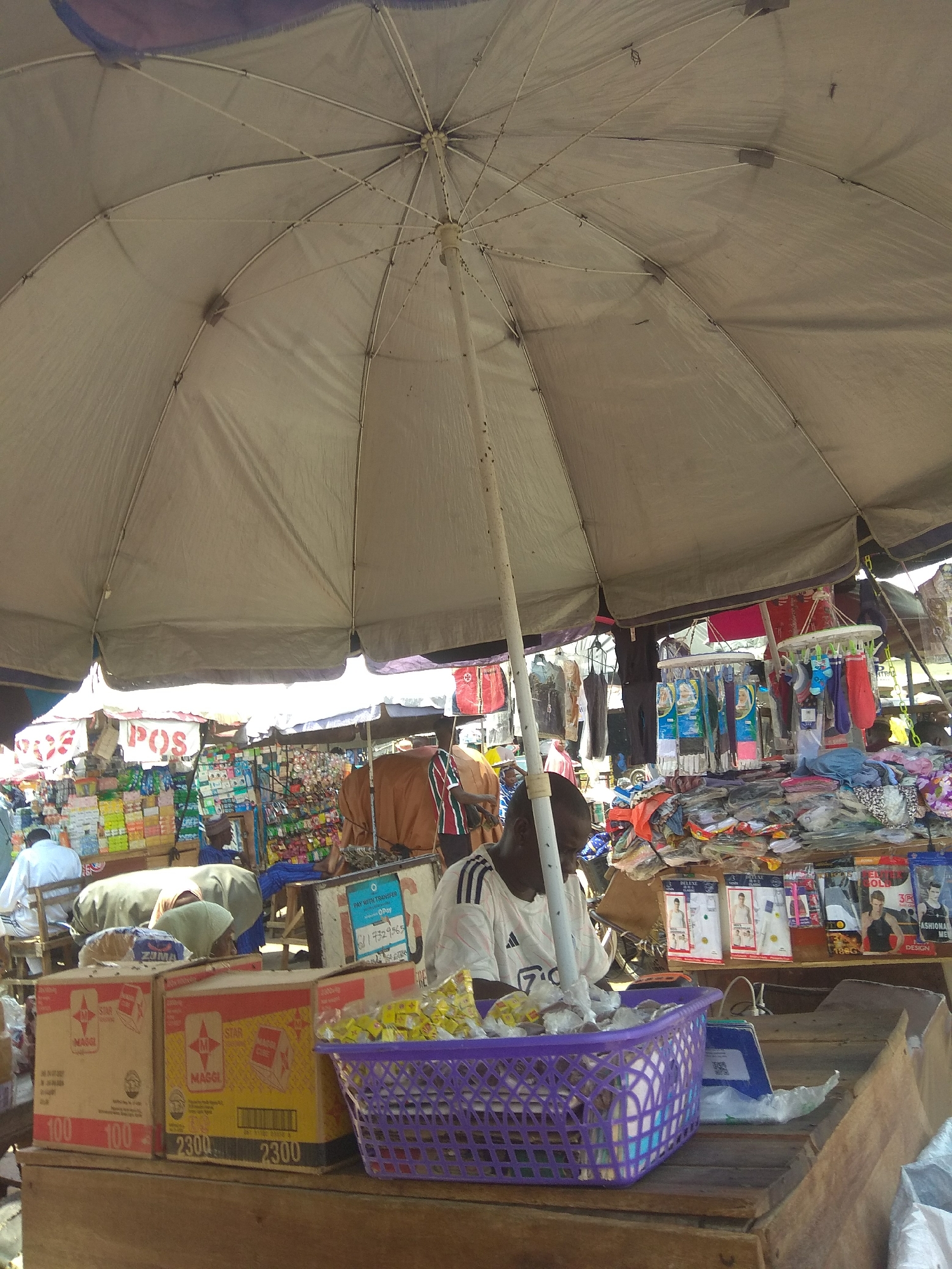 This media file depicts a bustling market scene in Jalingo, Nigeria. The primary subject is a vendor sitting behind a wooden stall, sheltered by a large, pale-colored umbrella. He is wearing a white t-shirt with black stripes on the sleeves and appears to be attending to his merchandise, which includes a purple basket filled with various items, some of which are packaged in plastic.
In the background, the market extends outwards with stalls displaying a variety of goods, including clothing, packaged food items, and other miscellaneous products. Several other people are visible in the scene, some appearing to be customers browsing the wares or fellow vendors. The atmosphere suggests a typical daytime market activity, likely under a bright, sunny sky, given the strong shadows cast by the umbrella and structures.
Visible text in the image includes branding like "MAGGI" and "STAR" on cardboard boxes, indicating food products. Other signs, such as "POS" and "OPay" with transaction details, point to payment and business operations within the market. The overall impression is one of a vibrant and active commercial hub in Nigeria.