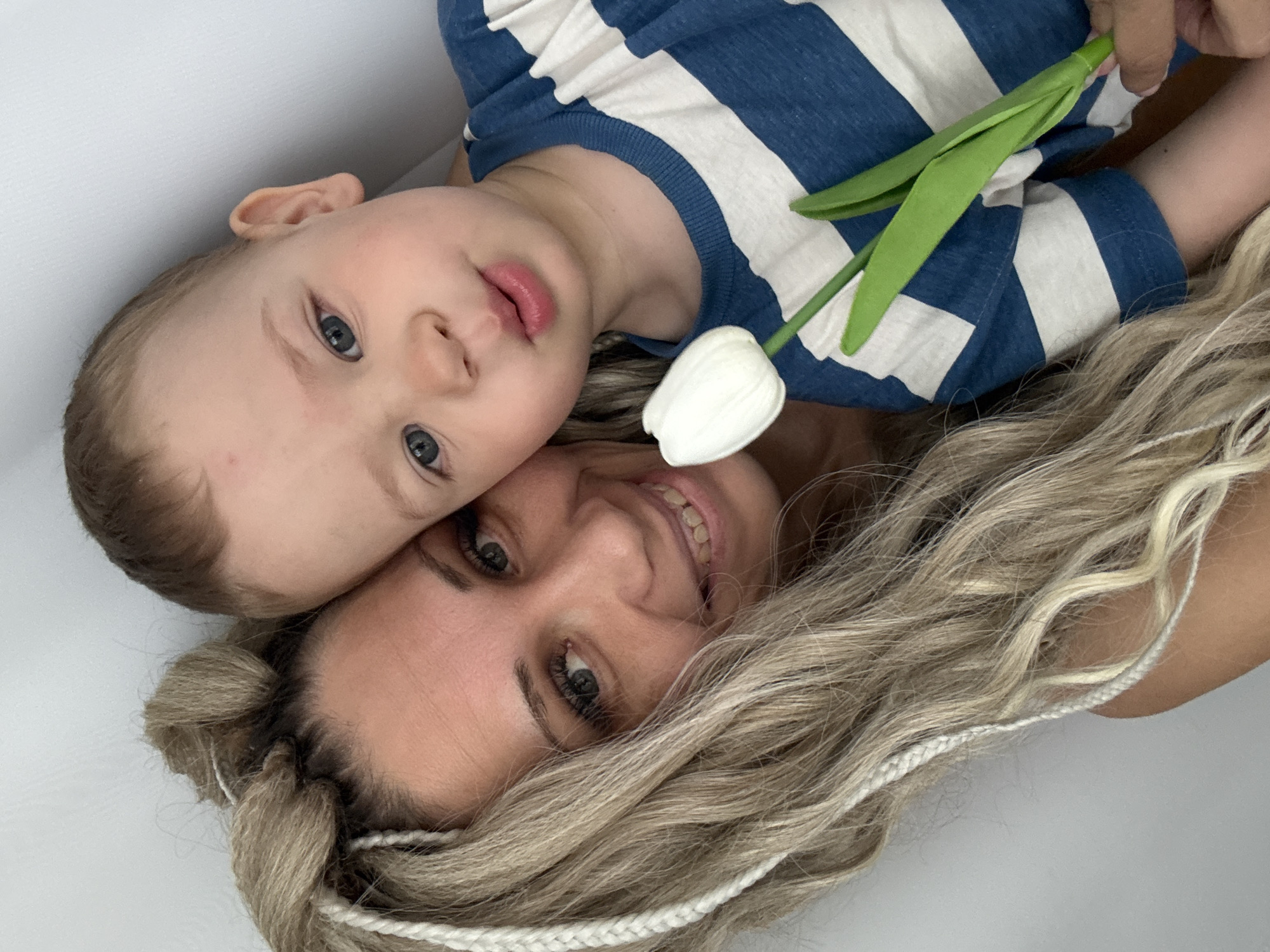 A woman and a young child are pictured in a close-up shot. The child, wearing a blue and white striped shirt, is lying on the woman. The woman has light blonde hair with white braids and is smiling. A white tulip flower is positioned between them. The location appears to be indoors and is unknown if it is Rusanovo, Russia.