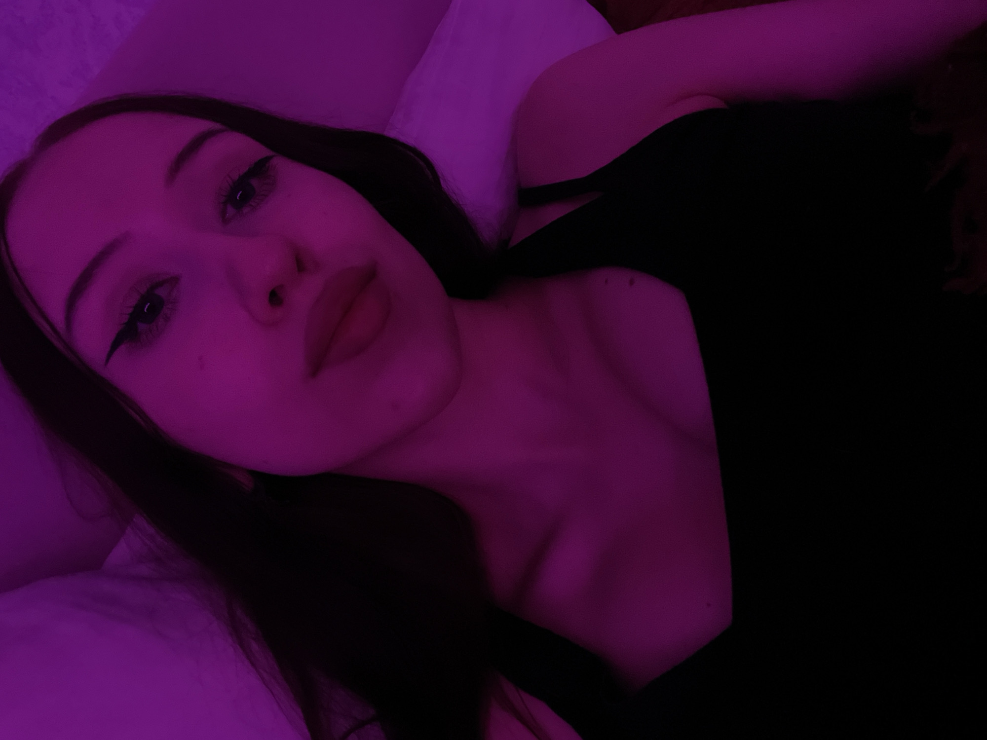 A young woman with dark hair and makeup is shown from the chest up, illuminated by purple lighting. She is wearing a black top. Her head is tilted slightly to the left, and she is looking directly at the camera. The background consists of a textured surface, possibly a wall or bedding, also in shades of purple. There is no visible indication of her location in Votkinsk, Russia, or any other specific geographical context.