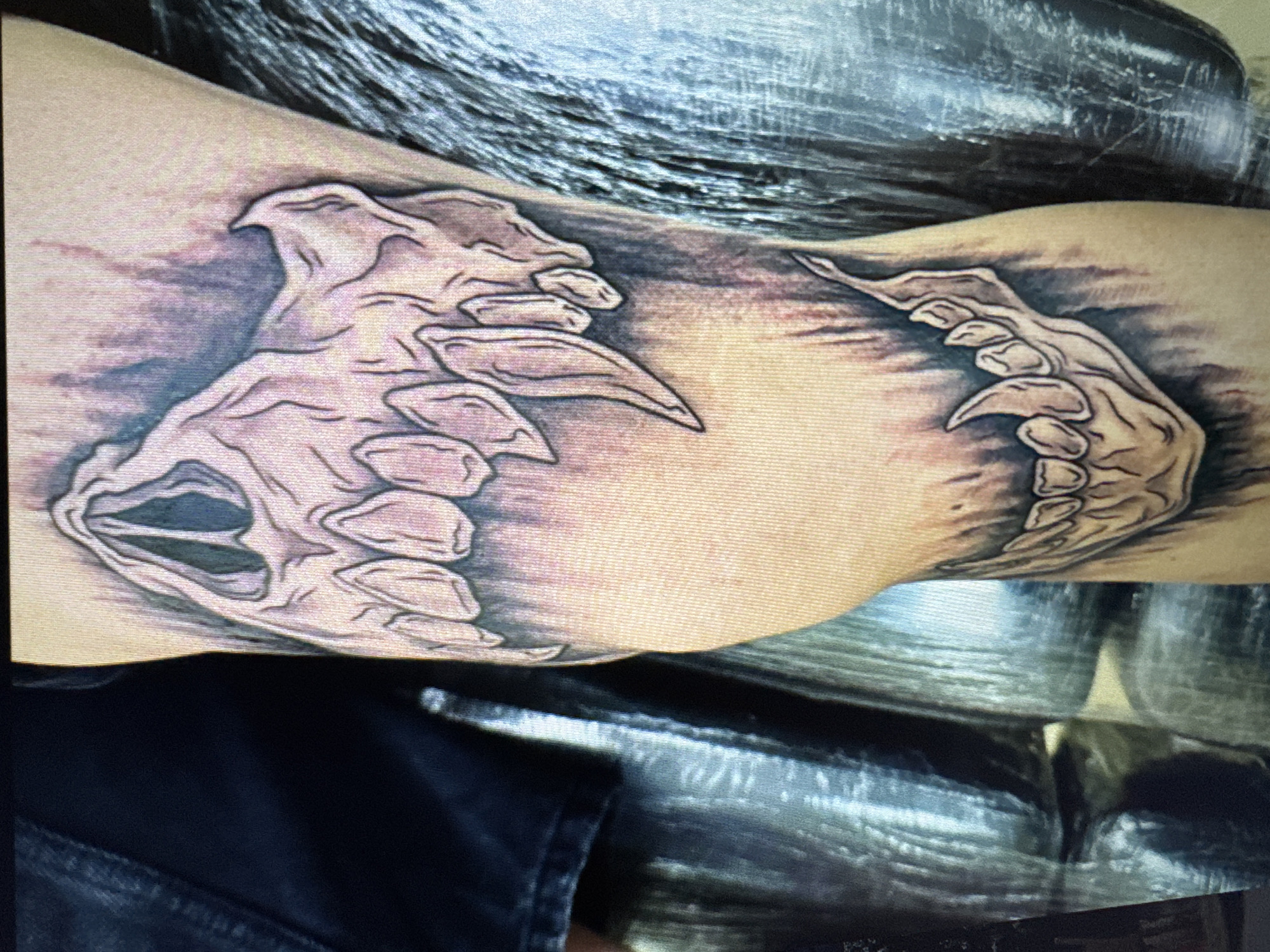 The image shows a close-up of a tattoo on a person's leg. The tattoo depicts a skeletal jaw with sharp teeth and is rendered in shades of black, grey, and pale pink. The background of the tattoo has a smoky or distressed effect. The leg appears to be that of an adult, and the skin tone is light. A dark blue garment, possibly denim, is visible on the left side of the frame. To the right of the leg, a textured, reflective surface is present, which could be a piece of furniture or a wall covering. There is no discernible indication of the city of Varna, Bulgaria, or any specific urban environment within the image.