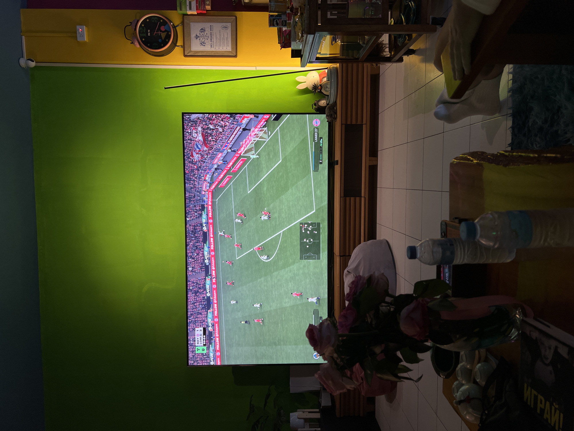 The image shows a living room or entertainment space, likely inside a home. A large television screen dominates the scene, displaying a soccer game, possibly a FIFA video game, featuring the teams BAY (Bayern Munich) and LOM. The screen is centrally located on a wooden console. A section of the wall behind the television is painted in a bright green color, with a darker tone visible on the side. The wall decoration includes items such as a clock, framed art, and a CCTV camera.
The foreground includes a bouquet of pink roses in a vase, along with several water bottles and a partially visible newspaper with the word "ИГРАЙ!" (play). In the top right corner, a person is visible using a cleaning sponge. The room is tiled, and there are objects placed on a stand or table in the background, possibly decorations or household items. The OCR results show text on the television screen, and the word "ИГРАЙ!" from the newspaper.