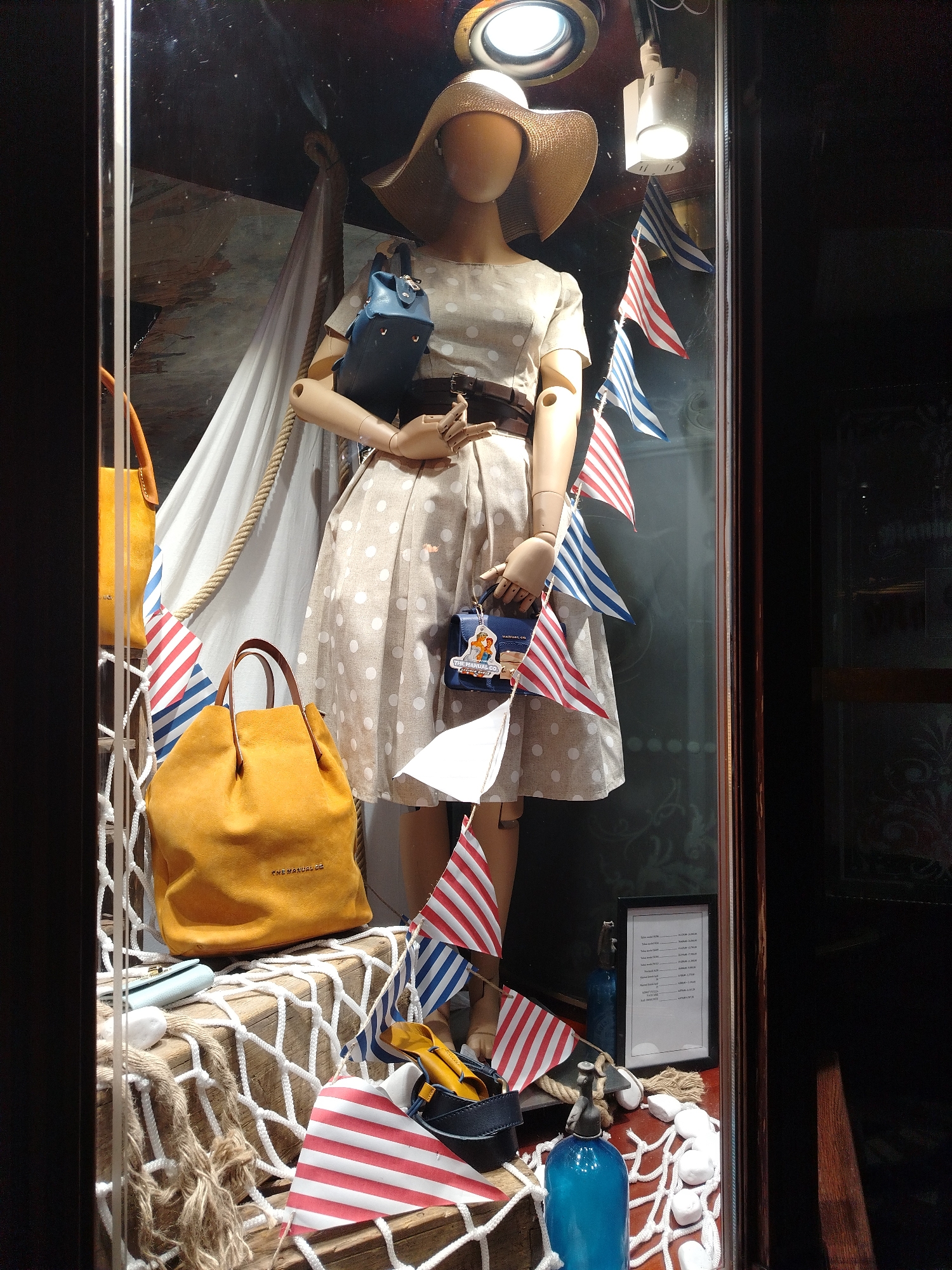 The image is a photograph of a shop window display, likely in Novi Sad, Serbia. The display features a female mannequin dressed in a polka-dot dress, a wide-brimmed straw hat, and a brown belt. She is accessorized with two handbags, one large mustard yellow tote bag and a smaller navy blue crossbody bag. The mannequin is positioned behind a wooden structure decorated with a fishing net, ropes, and various nautical-themed items, including small flags with red and white stripes, and blue and white stripes. To the left of the mannequin, another mustard yellow tote bag is visible. In the foreground, there are additional decorative elements like white stones, a blue glass bottle, and a framed price list. The overall theme of the display appears to be nautical or summery.