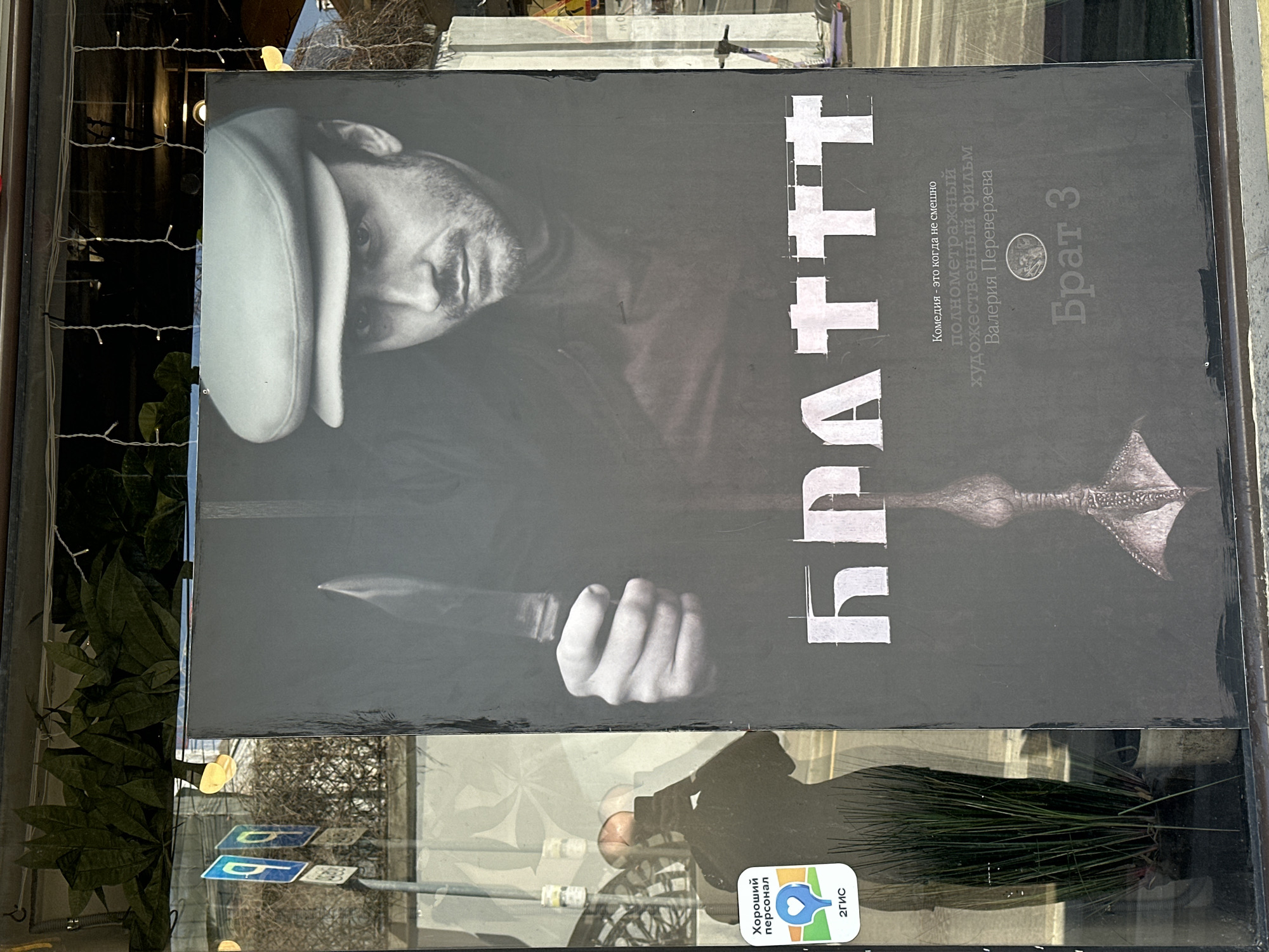 A poster of the Russian film "Brat 3" (Brother 3) is displayed in the window of a store. The poster features a man wearing a hat, holding a knife. The name of the movie is written in large, white Cyrillic letters. There is also some text in smaller print describing the film as a comedy. A 2GIS "Good Staff" sticker is attached to the glass. Reflected in the window are street signs, a bush, and parts of a person. The location is Moscow, Russia.