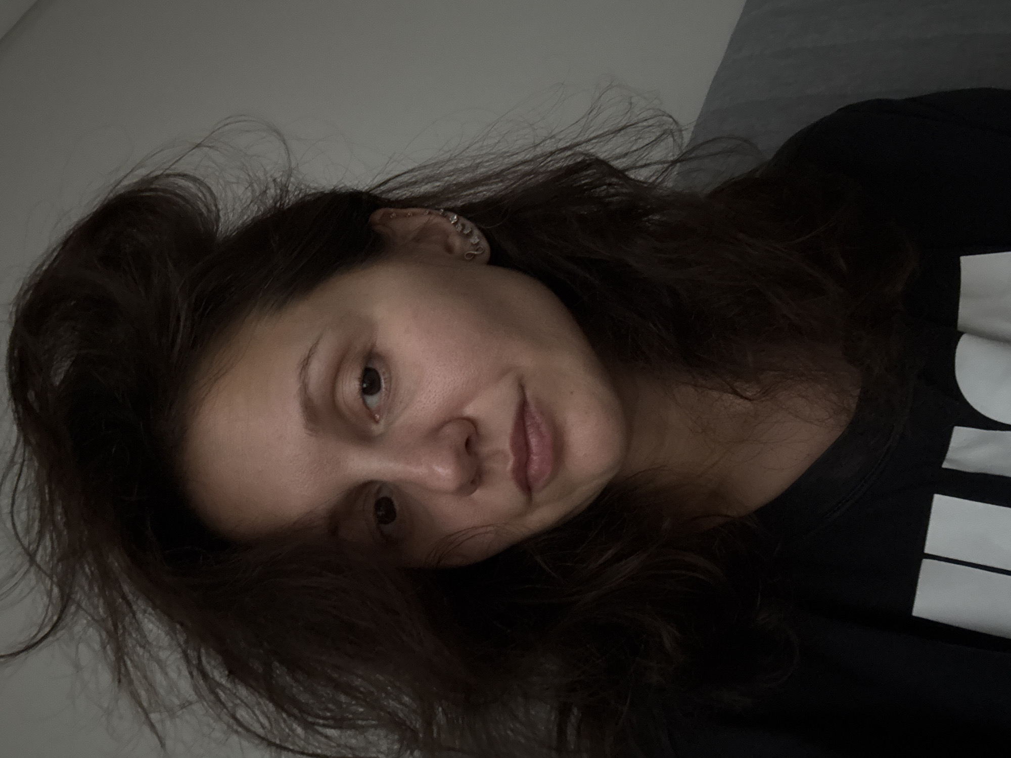 A close-up, angled portrait features a young woman with long, dark, wavy hair, which appears somewhat disheveled. Her head is tilted to the right, and she looks directly towards the viewer with a soft, gentle expression, hinting at a slight smile. Her skin is clear, and her eyes are dark. Her left ear, partially visible through her hair, has multiple piercings, including a distinctive silver-toned cuff or chain-like earring along the upper cartilage. She is wearing a dark, likely black, t-shirt, on which partially visible white block lettering can be seen on the right side of her chest. The background is a plain, light gray or off-white wall, suggesting an indoor setting. The lighting is soft and even, without harsh shadows. The overall mood is casual and relaxed. While the provided context indicates Dubai, United Arab Emirates, no specific visual cues within the image confirm this location.