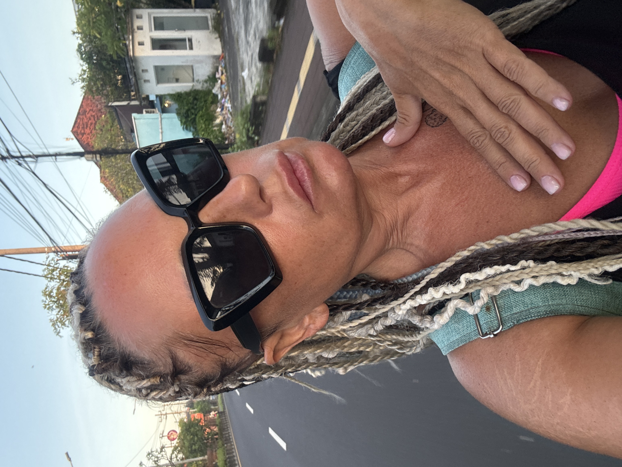 A selfie-style photograph features a light-skinned person with a close-up view of their face and upper body. The person is wearing large, black sunglasses. The individual's hair is styled in long, braided strands of varying shades of brown and white. A hand with light-colored fingernails rests on their upper chest/shoulder area. The person is wearing a black top with a hint of a bright pink undershirt visible. A light blue denim top is partially visible beneath the braids.
The background shows an urban environment, including a road with white lane markings. In the distance, there are buildings and trees. The location is Kuta Selatan, Indonesia. A small building with white walls and windows is situated in the background, surrounded by some foliage. Power lines are visible against the sky.