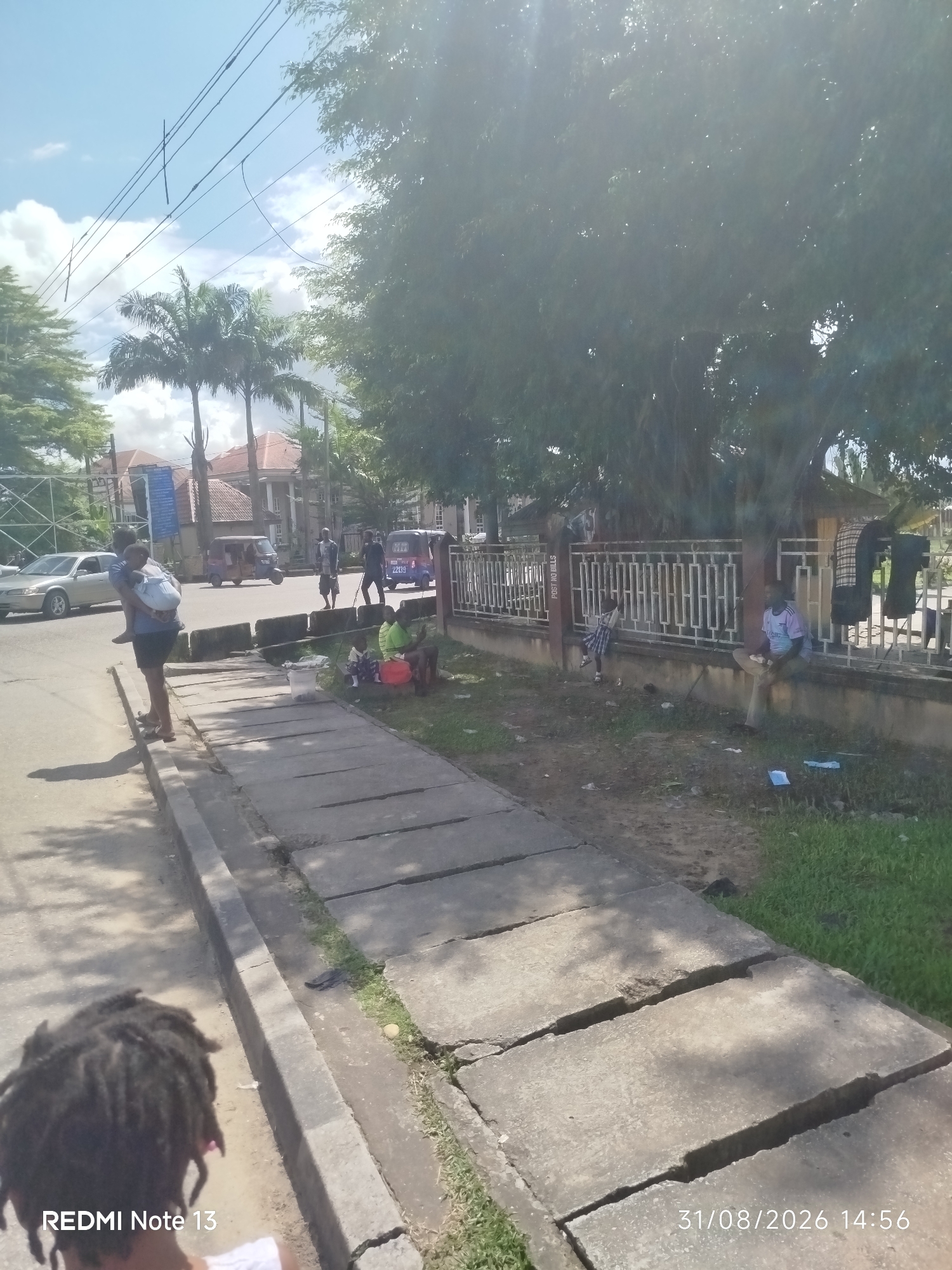 The image depicts a street scene in Yenegoa, Nigeria, under a bright, partly cloudy sky. In the foreground, a person with dreadlocks is partially visible from behind, facing away from the camera. To the left of the road, a sidewalk with cracked concrete slabs runs along the side. A woman carrying a child on her back is walking on the sidewalk. Further down the street, a silver car is parked, and a woman is standing next to it. Several people are gathered on the side of the road, including a group sitting on the ground, some appearing to be selling or interacting. A man is sitting on a low wall in front of a decorative metal fence. Behind the fence, clothing is hung out to dry. The background shows buildings and palm trees, typical of the Yenegoa landscape. The overall impression is a bustling urban environment on a sunny day.