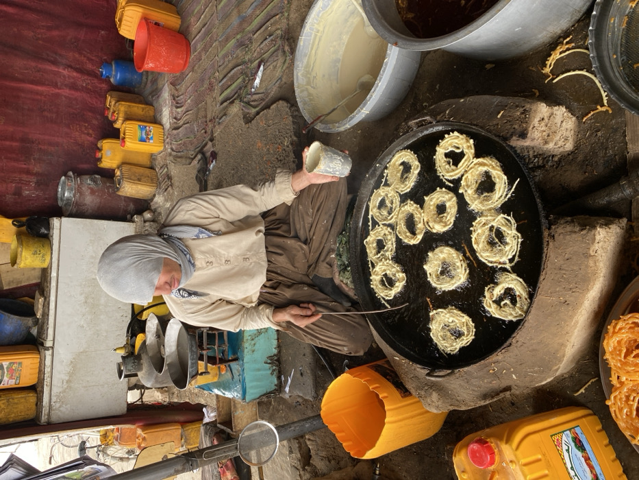 A person is depicted in a crouched position, actively frying ring-shaped sweets, consistent with Jalebi, at a street food stall in Kandahar, Afghanistan. The individual wears a light tan, long-sleeved shirt, dark brown pants, and a light grey head covering that partially obscures the forehead, with a patterned scarf visible around the neck. They manipulate approximately twelve light-golden, spiraled dough rings, which are sizzling in a large, dark, shallow pan filled with hot oil. The pan rests on a low, dark-colored, possibly earthen or brick base. The person uses a long, slender metal tool in their left hand and holds a small, grey, cylindrical cup in their right hand.
To the immediate right of the frying pan, a large, grey plastic basin contains a light-yellow batter, with a white ladle resting inside. Adjacent to this, a large metal basin holds a dark, reddish-brown liquid, indicative of syrup. In the lower left foreground, a stack of finished, syrup-soaked Jalebi rests on a metal plate. Scattered throughout the scene are numerous plastic containers: several yellow jerrycans of varying sizes, an orange plastic bucket on the left, and a red plastic bucket along with additional yellow and blue jerrycans in the background.
The background shows a makeshift environment with a red fabric or tarp wall. A white, chest-style cooler or refrigerator is partially visible behind the cook. Various other cooking implements, including pots and a gas burner, are present. The ground is unpaved, appearing as rough earth or dirt. A bicycle frame is partially visible on the far left side of the scene.