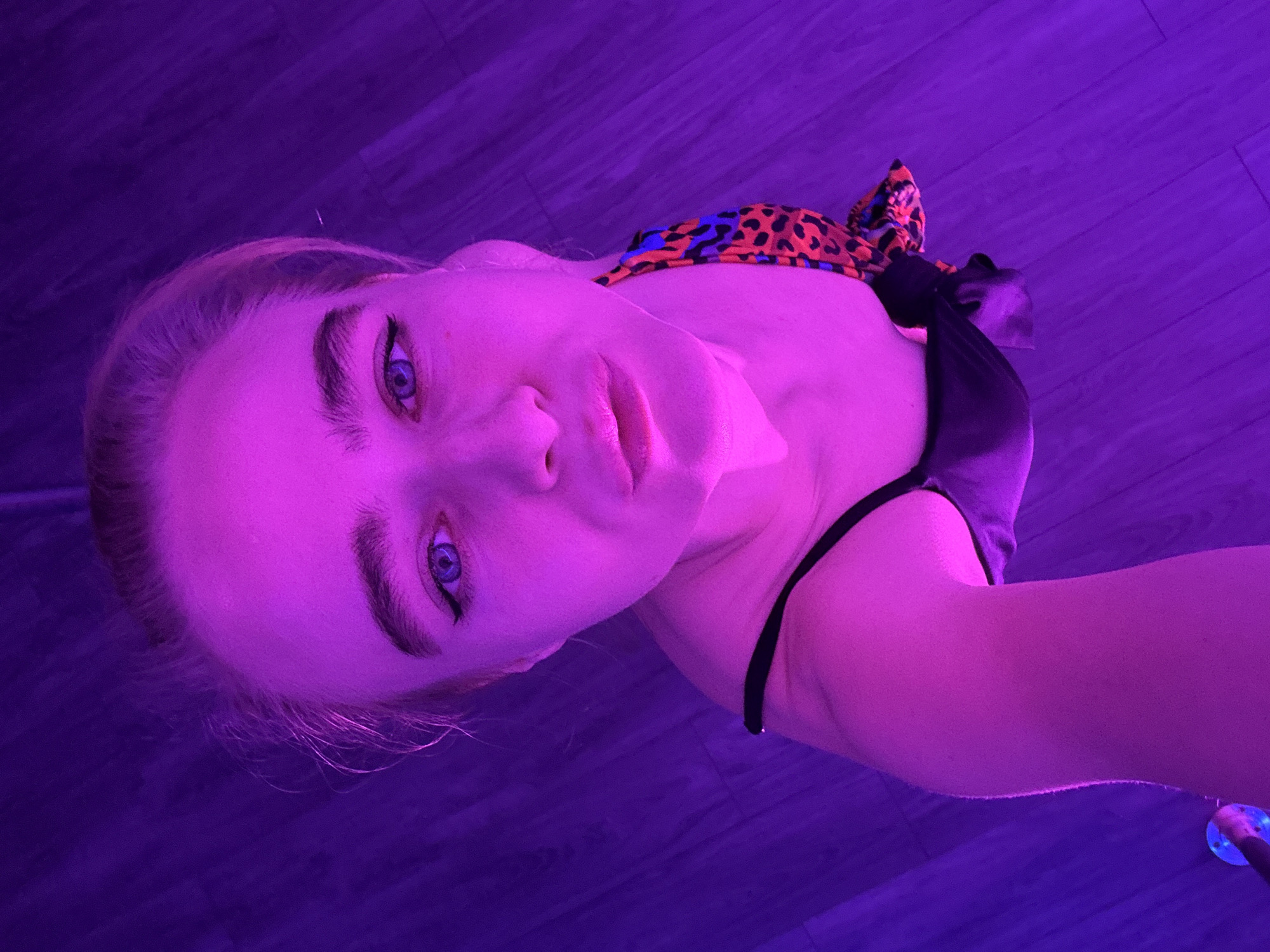 A young woman poses for a selfie with a purple hue cast over her face and surrounding environment. She has light skin, blue eyes, and is wearing a black top with an animal print scarf tied on top. The background appears to be a wood-look flooring, also bathed in purple light. The location is unknown, but presumed to be an indoor setting, possibly a studio or home, within the city of Unknown, Unknown.