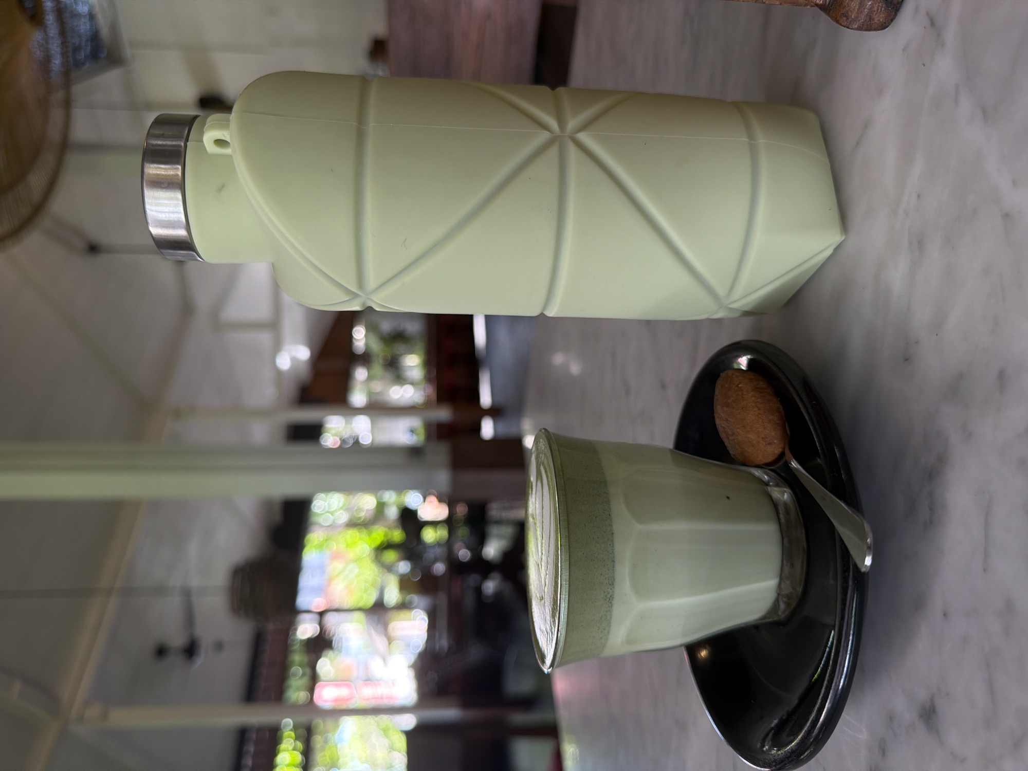 On a marble surface, likely a table, lies a light green water bottle with a geometric design and a stainless-steel cap. Next to it is a glass of a pale green liquid, possibly matcha latte, sitting on a black saucer with a spoon and a small, round cookie or pastry. The background shows an open, airy space with glimpses of foliage and a ceiling fan, indicative of an outdoor or semi-outdoor setting. The image may be taken in North Kuta, Indonesia.