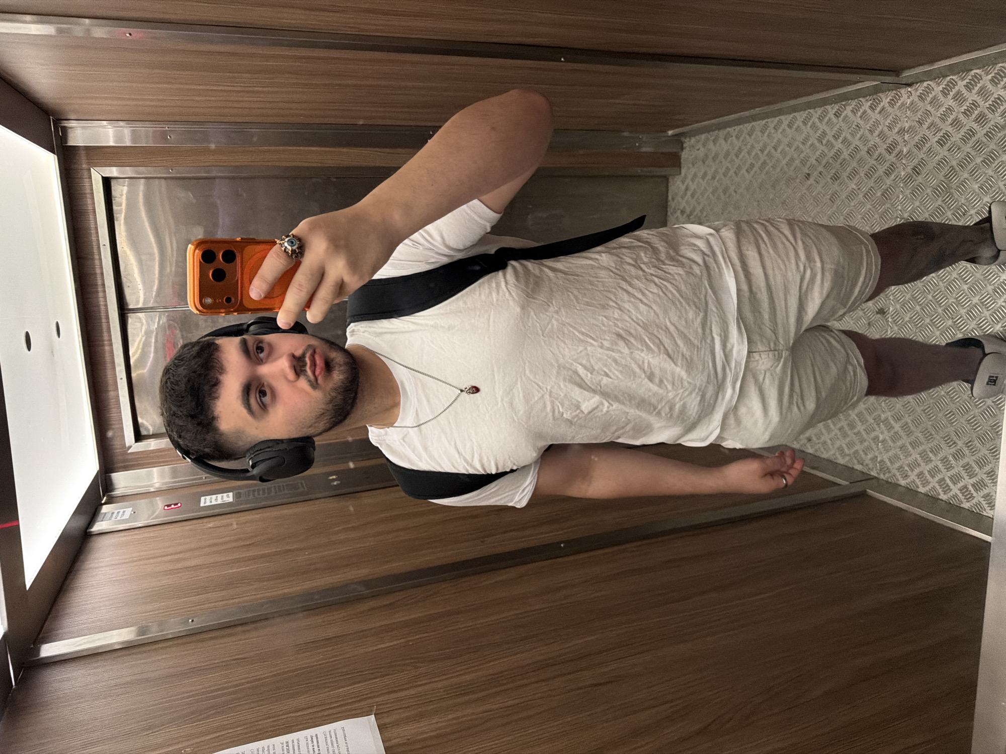 A young man with a beard and dark hair is standing in an elevator and taking a selfie. He is wearing black headphones, a white t-shirt, and light-colored shorts. He has a black backpack on his shoulders and a silver necklace with a red pendant. He is also wearing a ring on his right index finger and a watch on his left wrist. The elevator has wood-paneled walls and a metal floor. The man is holding an orange smartphone in his right hand to take the picture. The image is taken in Sofia, Bulgaria.