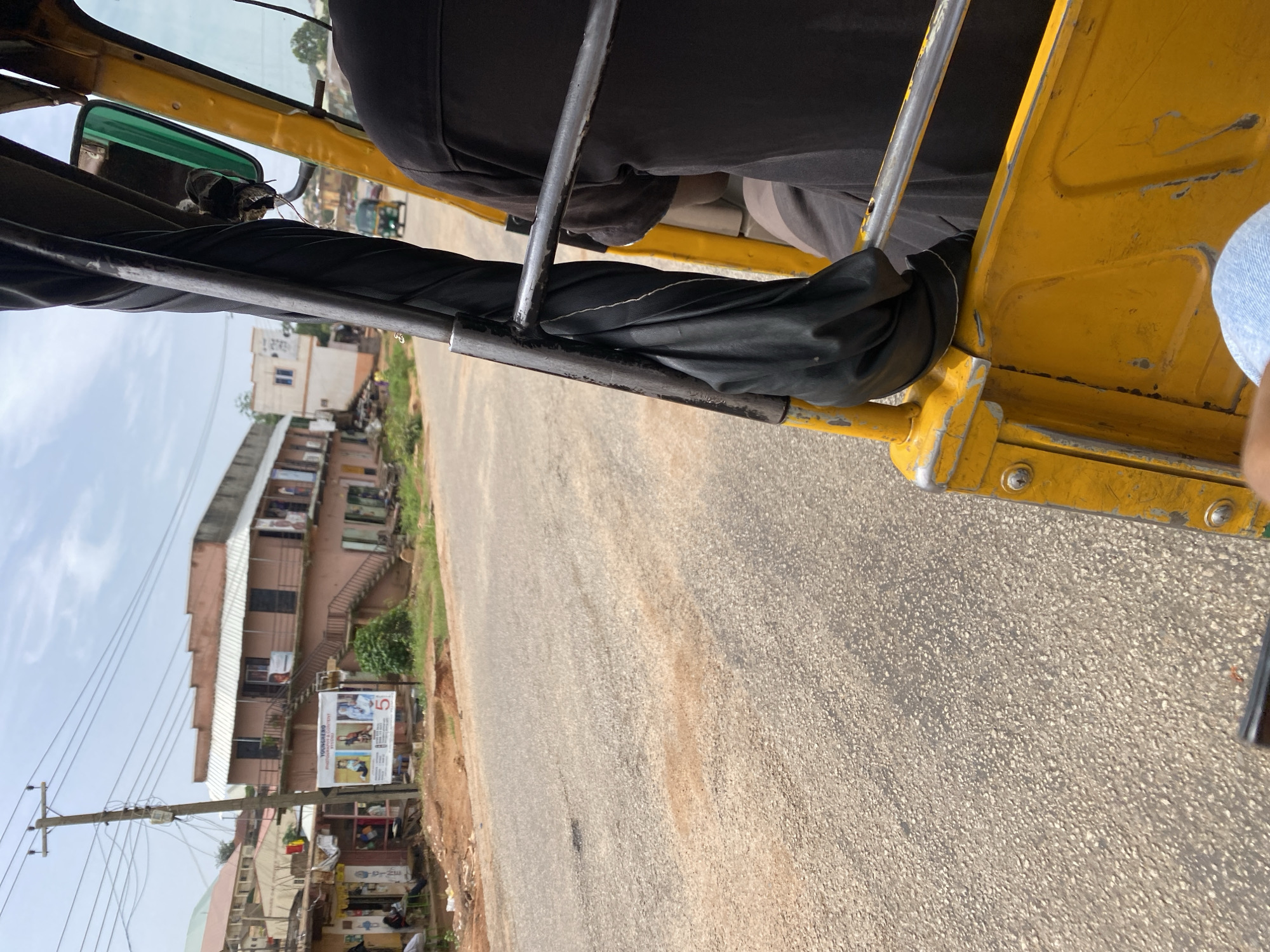 The image captures a view from the interior of a yellow auto-rickshaw, looking outwards at a street scene in Jos, Nigeria. The auto-rickshaw's frame and some of its interior, including a black cover and metal bars, are visible in the foreground. A person wearing a black shirt is seated in the auto-rickshaw.
The street is paved with asphalt and shows signs of wear, with patches of reddish-brown soil on the surface. To the left of the road, there are several buildings, some of which appear to be shops with awnings. A prominent billboard with images and text, including the word "YOUNGHERO" and the number "5", is displayed in front of one of the buildings. A utility pole with numerous wires stands on the left side of the street. In the background, more buildings and a glimpse of green vegetation can be seen. Another auto-rickshaw is visible further down the road. The sky is overcast with clouds.