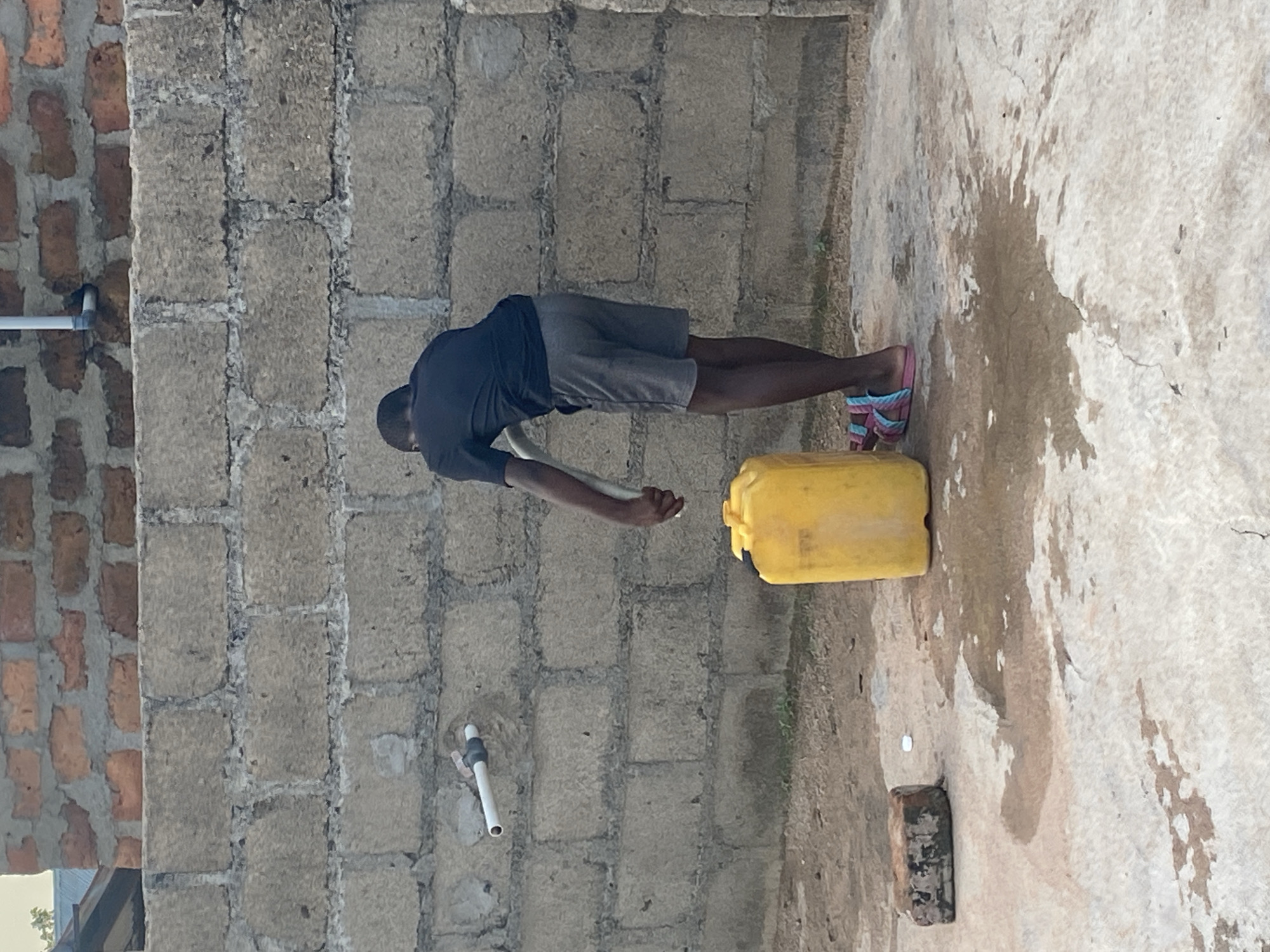 A person is standing in front of a cinder block wall, filling a yellow plastic jerrycan with water from a faucet. The person is wearing a dark t-shirt and grey shorts, with colorful sandals on their feet. The jerrycan is positioned on the ground next to a patch of wet concrete, indicating that some water has already spilled. A broken brick lies on the ground to the left of the jerrycan. The wall behind the person is made of rough-hewn cinder blocks, with a smaller section of red brick visible at the top. A white pipe emerges from the wall, connected to the faucet. The ground is a mixture of dirt and concrete.