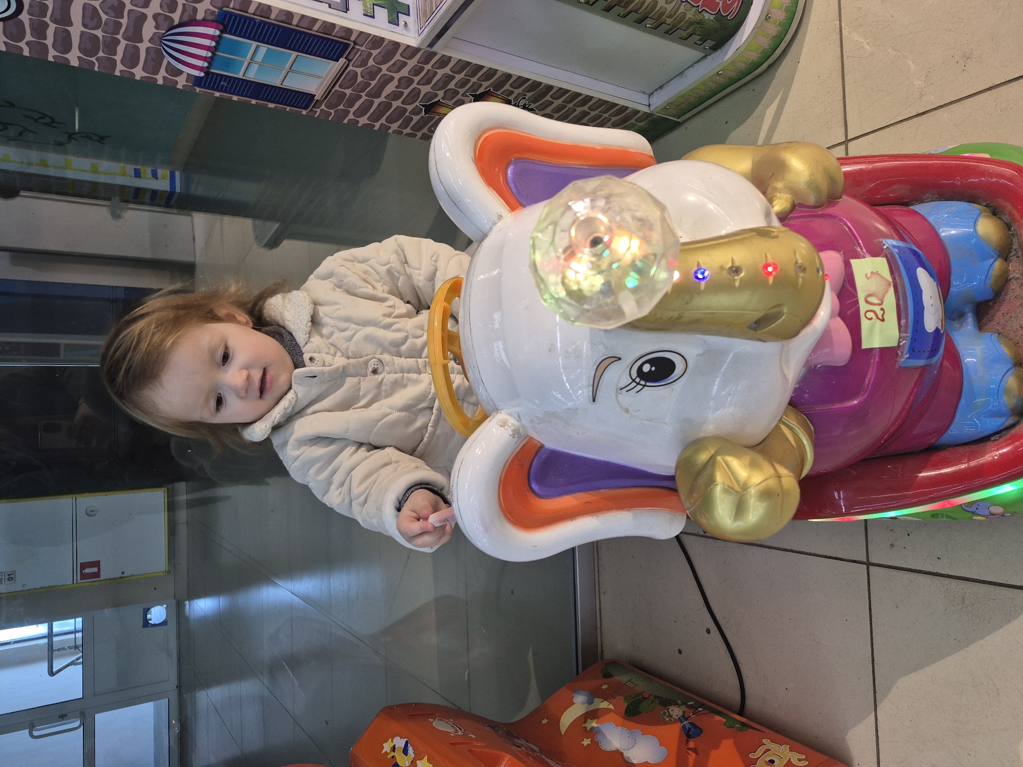 A young child is seen lying on the floor next to a coin-operated amusement ride in an indoor location. The ride features a stylized elephant design with a white head, large ears outlined in orange and purple, and gold-colored accents around the head and feet. The elephant has a plastic, multi-colored light fixture on top of its head, and its body is decorated in pink and blue. A yellow tag with the number 20 is attached to the side of the ride. The child is wearing a light-colored, quilted jacket and is positioned such that they are leaning against the side of the ride. The floor is tiled and reflective, showing a partial reflection of the ride and the child. In the background, there is a fire extinguisher cabinet, a wall with a tiled pattern, and a glimpse of an outdoor area with a door or window. A portion of another amusement ride, orange in color, is visible in the lower part of the frame. The city and specific indoor location are not discernible.
