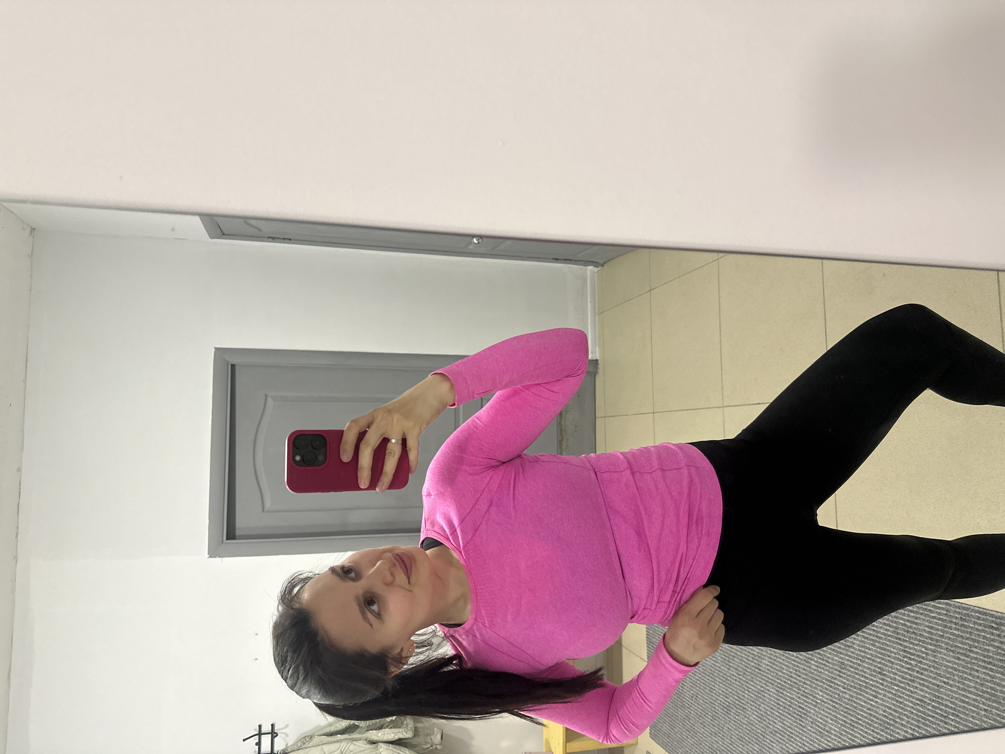 A woman is taking a selfie in a mirror. She is wearing a pink long-sleeved shirt and black leggings. She is holding a red phone in her right hand. The background appears to be a room with a gray door and light-colored tiled floor. The location is Chaikovsky, Russia.
