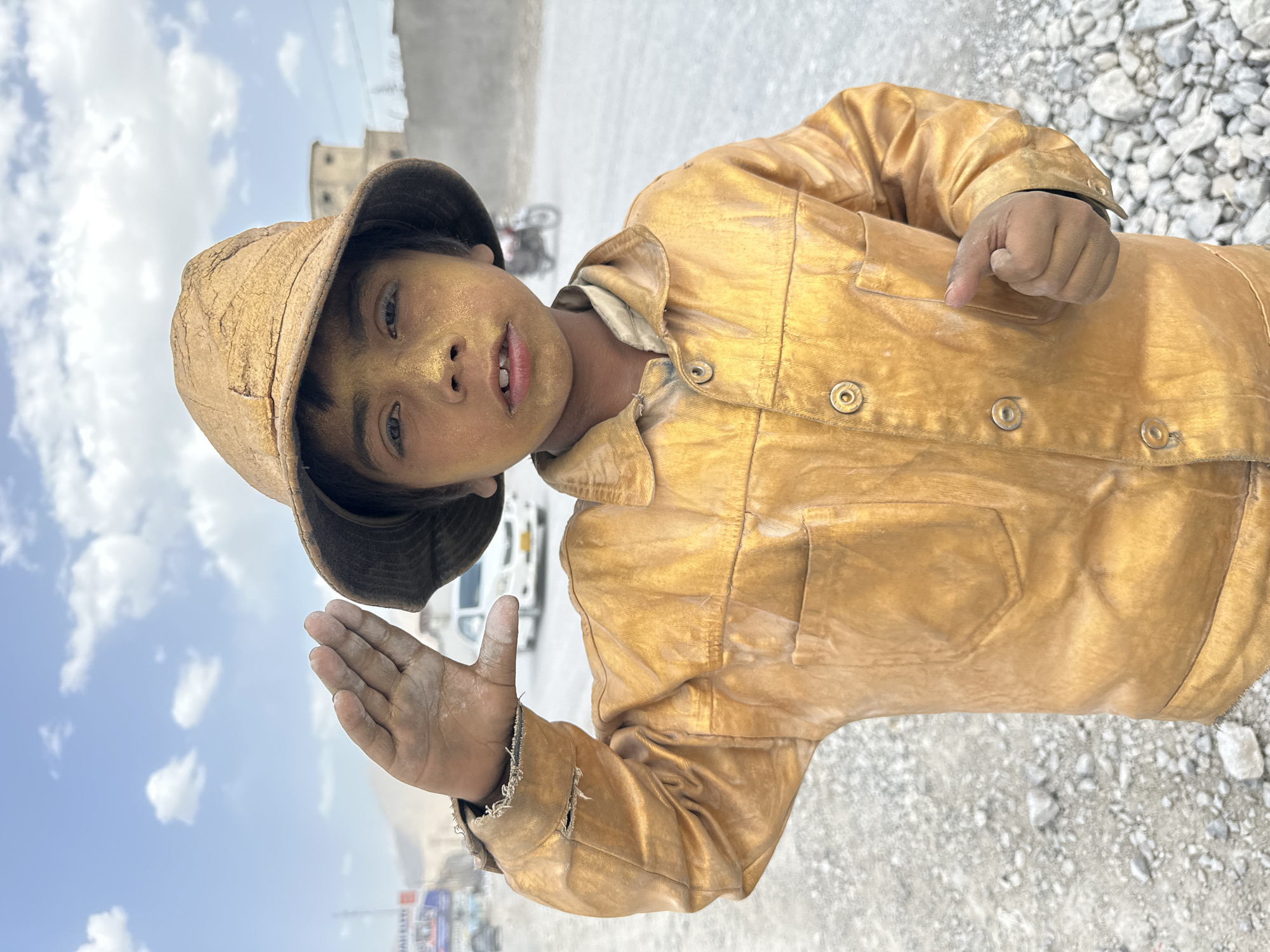 A young person with dusty skin and a hat is wearing a yellow jacket and is standing outdoors on a rocky surface. They are looking directly at the camera with one hand raised. A building and vehicles are visible in the background. The image was taken in Quetta City Tehsil, Pakistan.