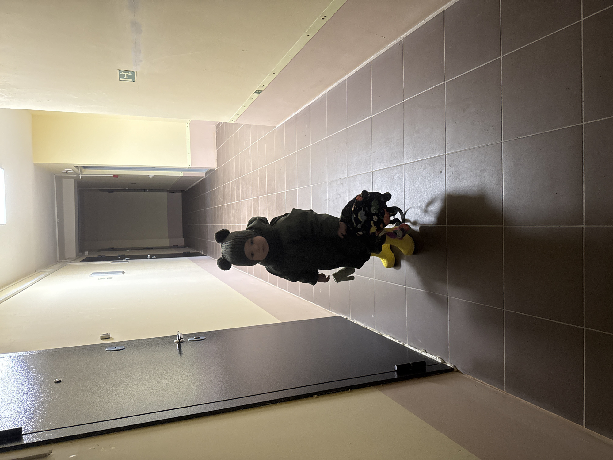 An indoor scene depicts a young child standing in a long corridor, viewed from a slightly elevated perspective looking down its length. The child, likely a toddler, is positioned roughly in the middle distance, facing the camera with a neutral expression. They are dressed in a dark, possibly black or dark green, padded jacket and a matching beanie with two pom-poms resembling bear ears. Bright yellow wellington boots are visible on their feet, and a small, colorful backpack with a dark base and various light-colored patterns is on their back.
The hallway features a tiled wall on the right, consisting of medium gray square tiles with white grout. The opposite wall on the left is painted a uniform light cream or pale yellow color, with a darker baseboard. The floor is composed of various materials, including light cream, a slightly darker beige strip, and a section of medium gray tiles that appear to match the wall tiles. In the immediate foreground, a dark, metallic-looking rectangular panel or door lies open on the floor, revealing several silver-colored latches or hinges.
Above the light-colored wall on the left, a large, rectangular, light-yellow structure is attached near the ceiling, possibly a decorative element or part of a ventilation system. The ceiling itself is a plain light cream color, and further down the corridor, a green emergency exit sign with a white running figure pictogram is visible on the ceiling. The lighting is artificial, suggesting an indoor setting, likely in an apartment building or similar public access facility in Chaikovsky, Russia, as per the provided context. The child appears to be paused in motion, perhaps exploring the hallway.