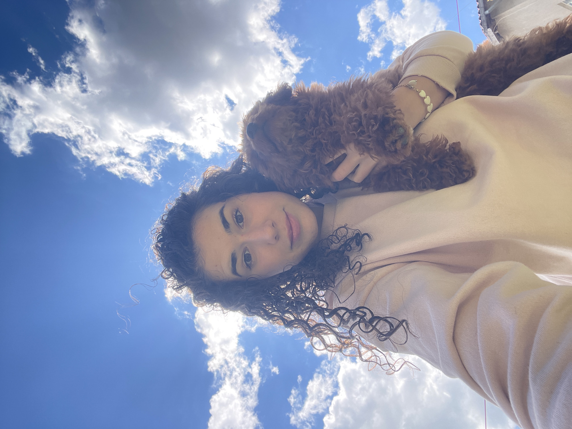 A selfie taken outdoors features a woman with curly hair and a brown poodle. The woman is wearing a light pink sweatshirt and a bracelet with heart-shaped charms. The dog is resting on her shoulder. The background includes a blue sky with white clouds. The location cannot be determined from the image, so city and country information are unverified.