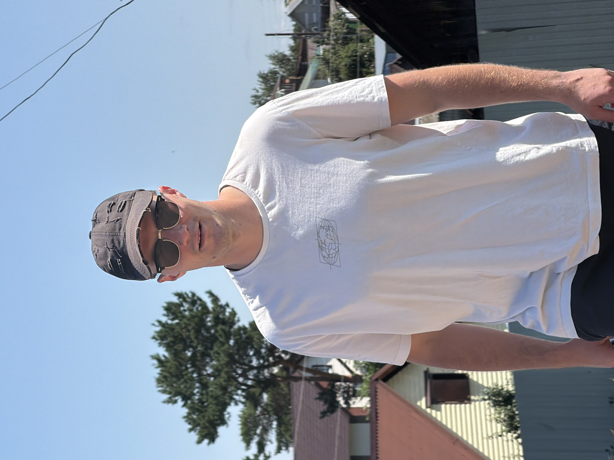 A young adult male is standing outdoors, looking towards the camera. He is wearing a dark baseball cap with rips and distressed detailing, sunglasses, and a plain white t-shirt with a subtle graphic design of a globe. The background features houses with visible roofs, trees, and a bright blue sky. The presence of houses and fences suggests a residential area.
