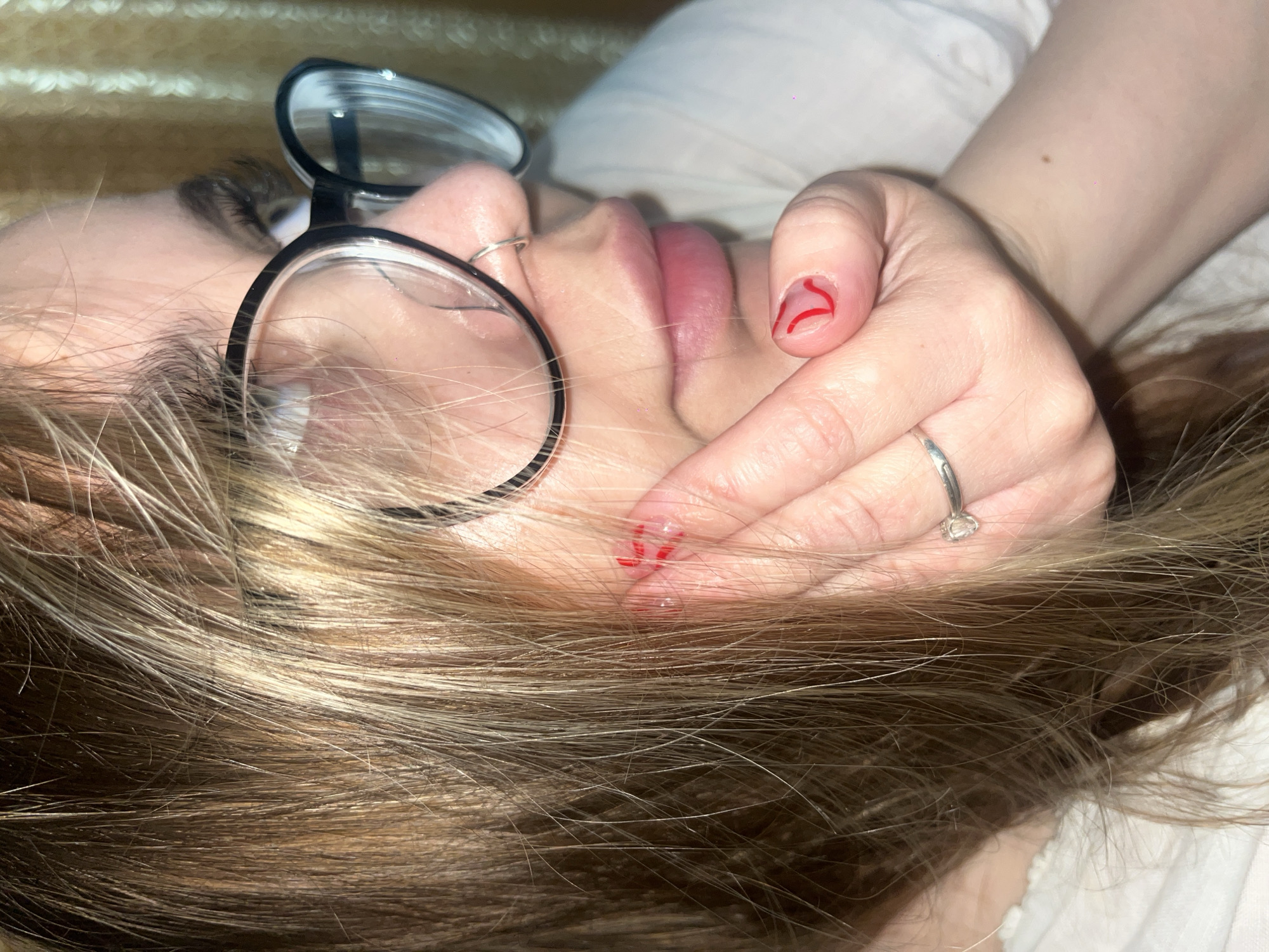 An eye-level close-up depicts a fair-skinned individual with light brown and blonde hair lying on a white surface. The person is wearing black-rimmed glasses and a silver ring on their left hand. Red, stylized lines are painted on the fingernails. The background is a blurred beige color. There is no evidence to indicate this image was taken in Novopokrovskaya, Russia.