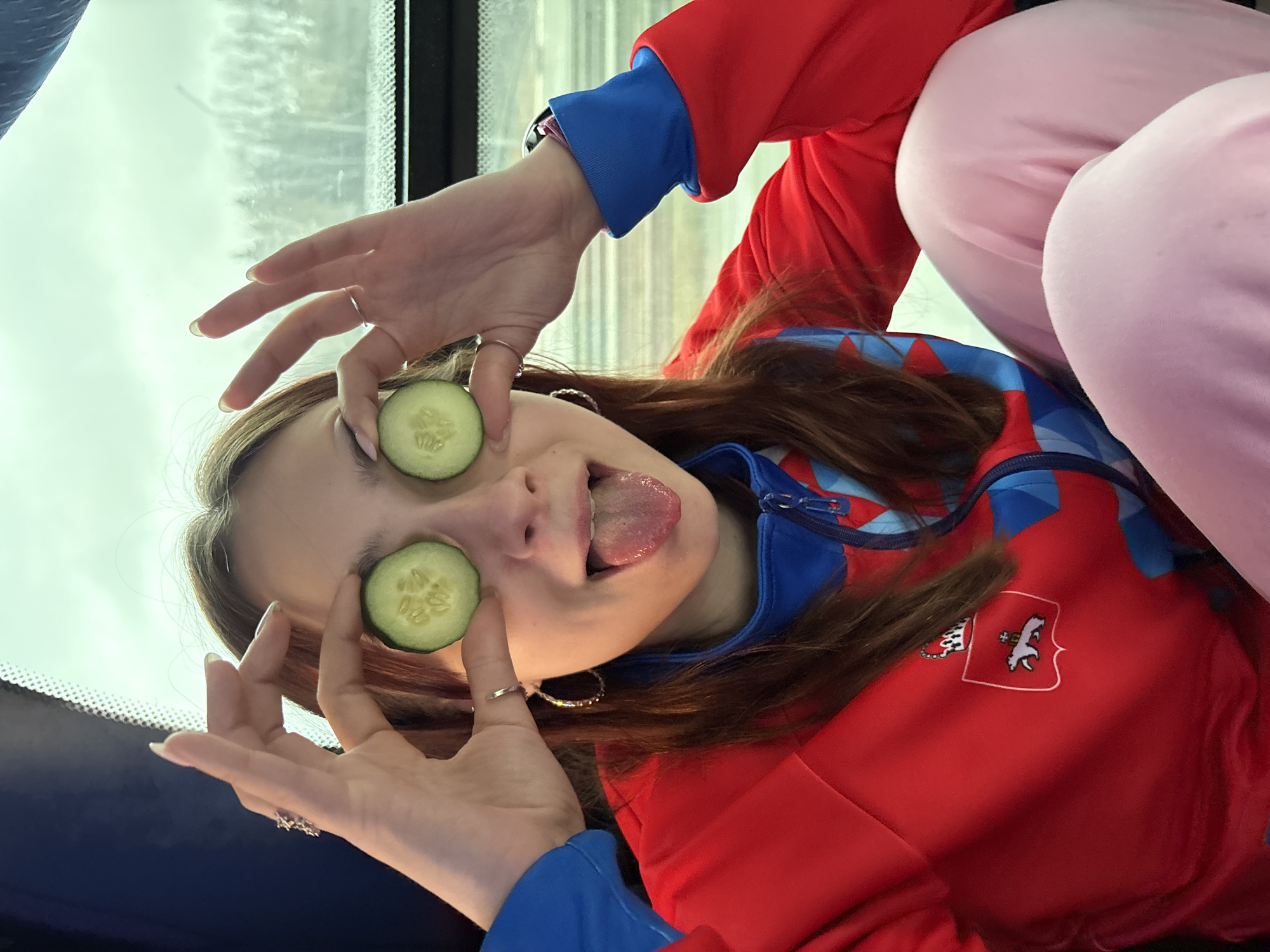 A young woman is holding cucumber slices over her eyes while sticking out her tongue. She is wearing a red and blue jacket, and pink pants. The emblem of the city of Yanychi, Russia, is visible on the jacket. The background appears to be outside a window.
