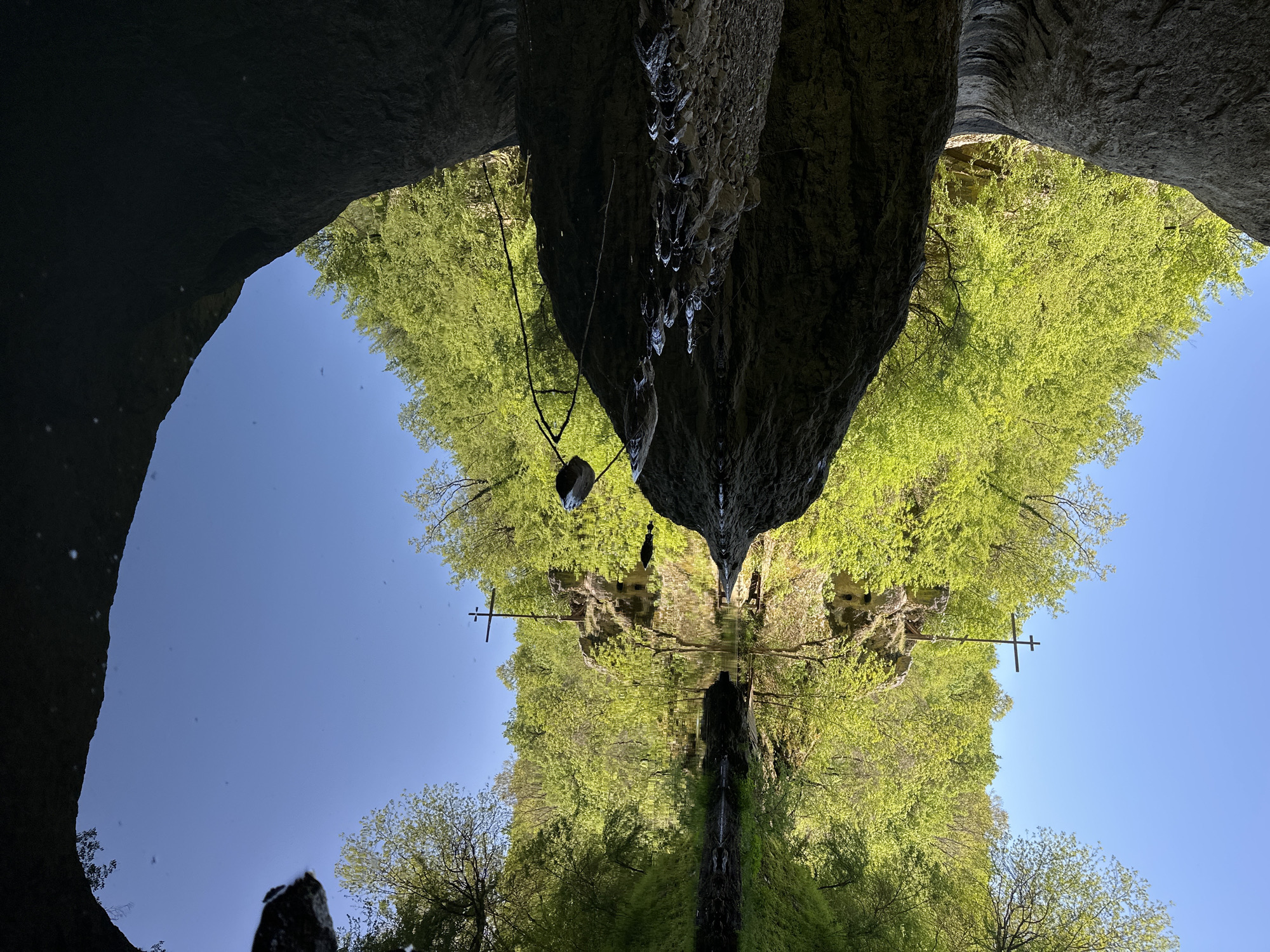 The image shows a reflection of a cave entrance with trees in the town of Lilyache, Bulgaria. The top of the image depicts a dark cave opening with water droplets falling from the rock face. The bottom half of the image shows a clear reflection in the water, mirroring the cave entrance, trees, and a clear blue sky. A metal cross is visible on the right side of the image, reflected in the water. The trees are lush green.