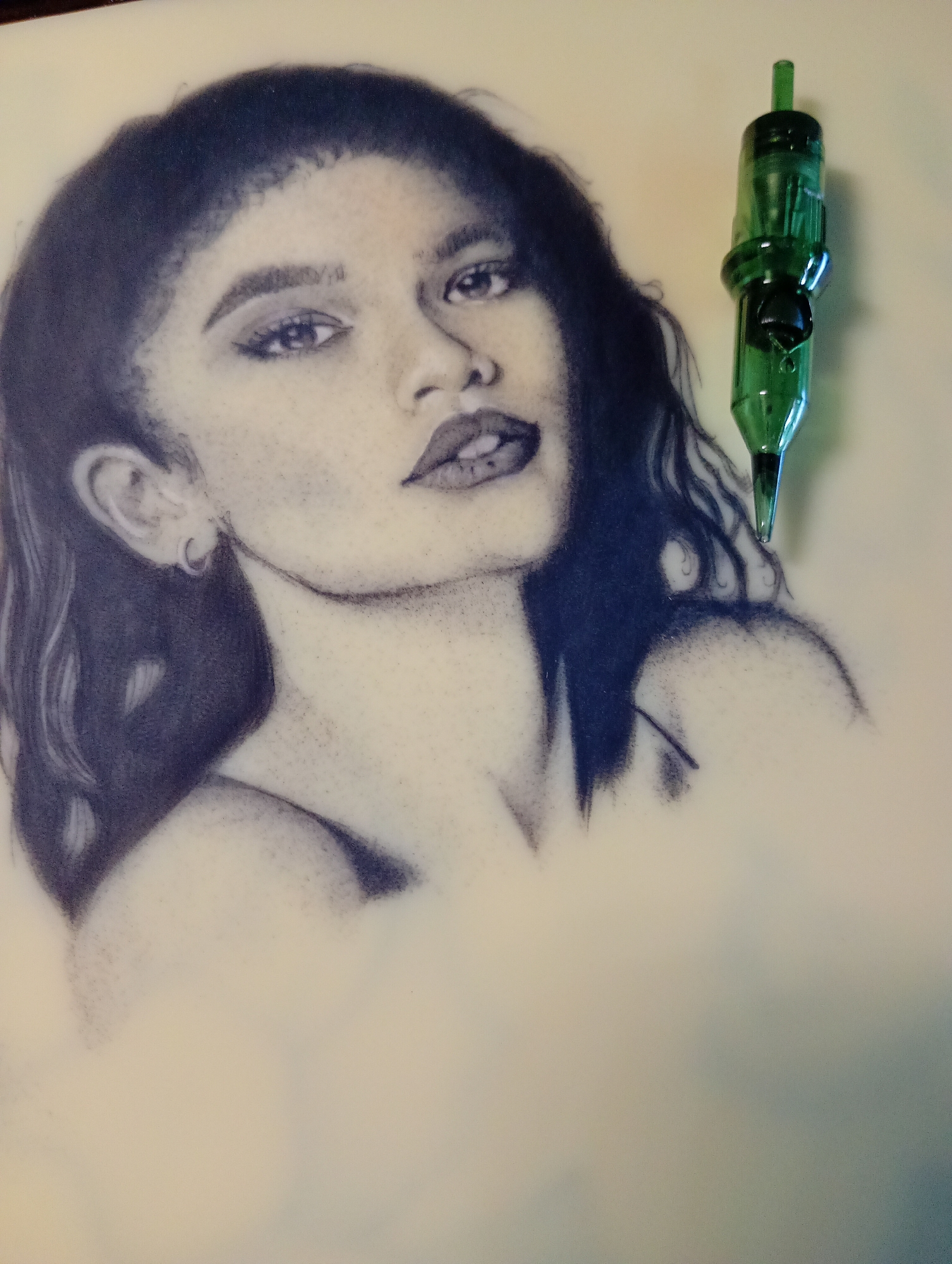 The image shows a drawing of a young woman with dark hair, dark eyes, and full lips. She is wearing small hoop earrings. The drawing appears to be done in a stippling style with blue ink on a white surface. To the right of the drawing is a green tattoo needle cartridge. The background is plain and out of focus. There is no indication of Roza, Bulgaria in the image.