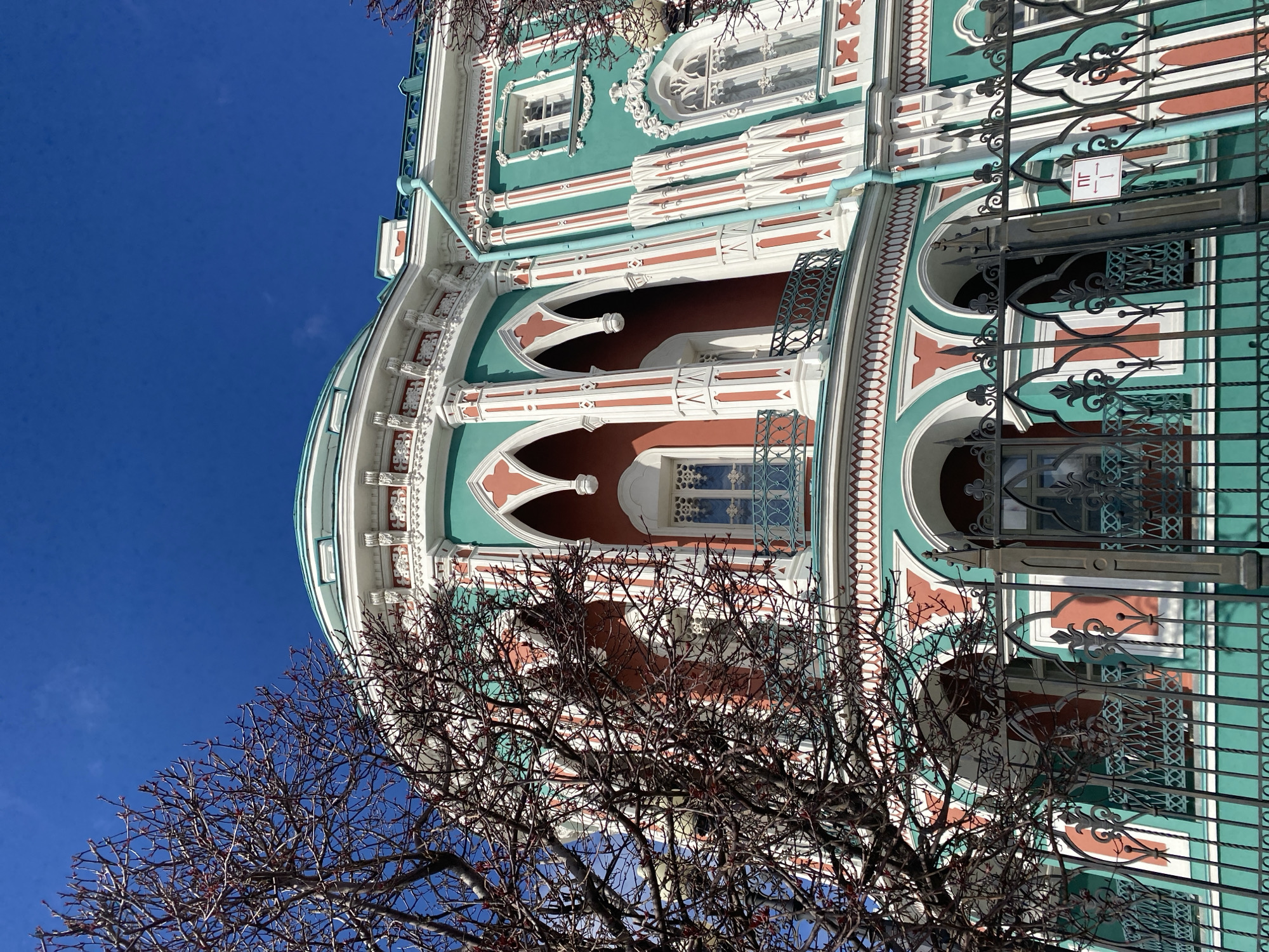 The image captures a section of the Sevastyanov House in Yekaterinburg, Russia, viewed from a low angle. The building showcases a distinctive architectural style featuring green and white facades with decorative red and white accents. The rounded corner of the building is prominent, adorned with arched windows and columns. A wrought-iron fence partially obscures the view of the lower portion of the building. A bare tree with budding branches is visible in the foreground. The sky is a clear blue. No people are visible in the scene.