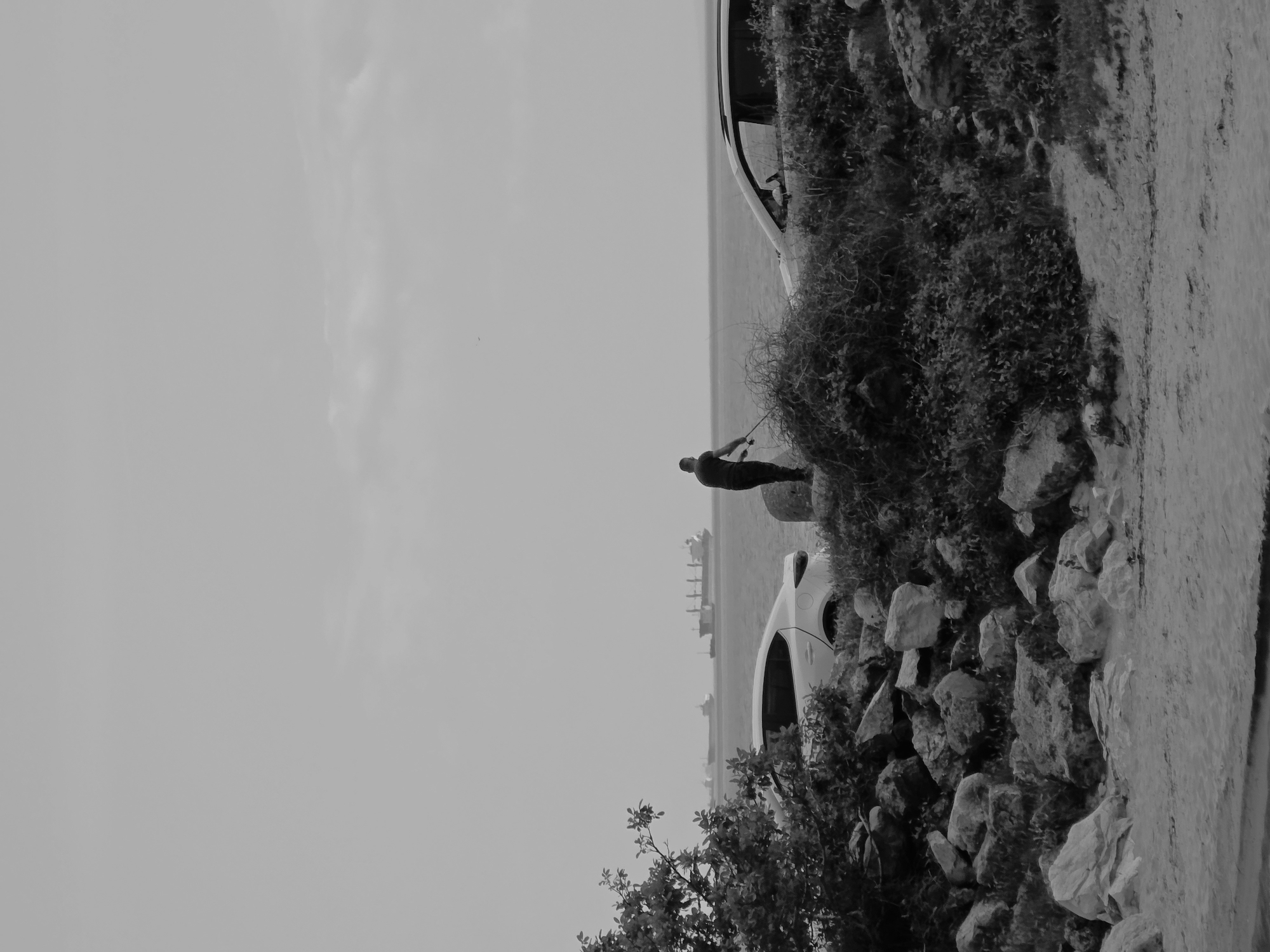 The image shows a scene in Varna, Bulgaria. A person is fishing on the coastline. A car is parked nearby. The scene includes rocks, vegetation, and the sea. The sky is overcast.