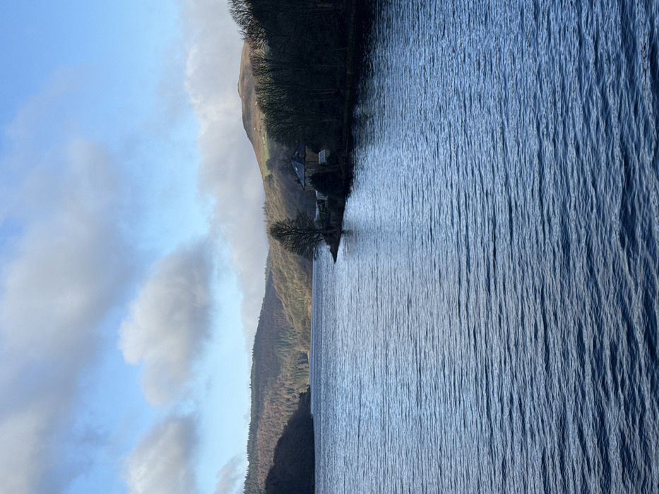 This image depicts Ladybower Reservoir, situated within the High Peak district of the United Kingdom, under clear daytime conditions. The foreground and midground are dominated by the dark blue, rippled surface of the reservoir water. Wooded hills, predominantly showing bare deciduous trees, rise from the water's edge on both the left and right sides, extending into the background. On the right side of the reservoir, a distinct promontory extends into the water, hosting a stone-built house with a dark grey or black roof, partially obscured by trees. The sky is bright blue with scattered white cumulus clouds, and sunlight is visible illuminating portions of the distant hills and clouds. No individuals or watercraft are present or discernible in the frame.