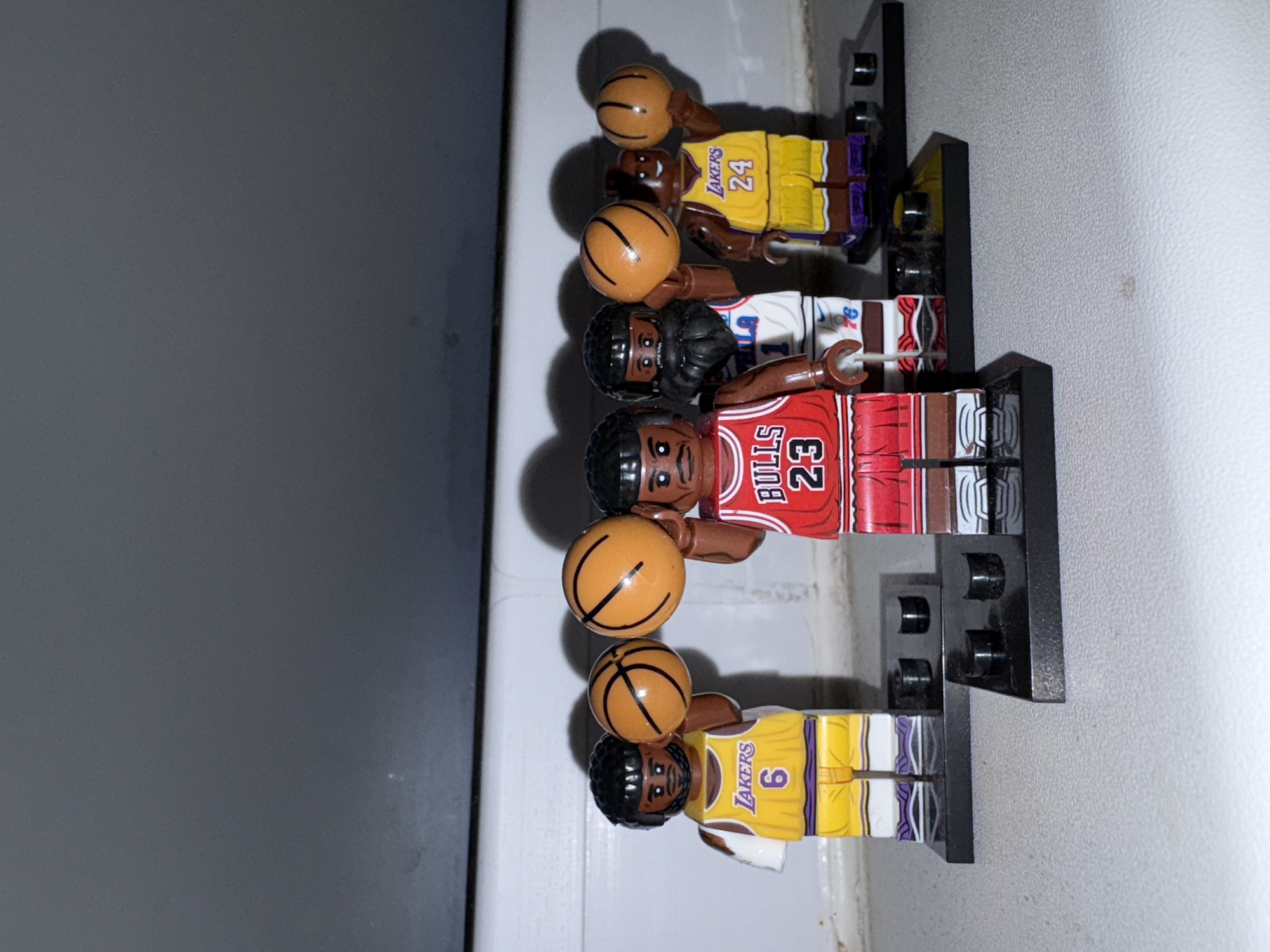 Four Lego basketball players stand in a row on black Lego stands. The first is wearing a yellow and purple Lakers jersey with the number 24. The second is wearing a white and blue Philadelphia jersey with the number 1. The third is wearing a red and white Bulls jersey with the number 23. The fourth is wearing a yellow and purple Lakers jersey with the number 6. Each figure is holding a basketball. The background is a gray wall. There is no indication of the city of Izhevsk, Russia.