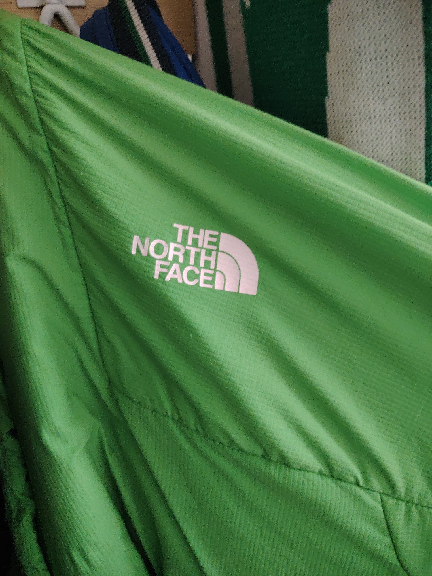 A close-up, angled photograph, taken in Huntingdonshire, United Kingdom, displays a vibrant lime-green outdoor jacket or windbreaker, characterized by a subtle ripstop grid pattern across its surface. The fabric exhibits various folds and wrinkles, suggesting it is draped or hanging. Prominently featured on the upper left section of the garment is the white "THE NORTH FACE" logo, partially cropped at the top. In the background, to the top left, a white plastic hanger hook is visible. Towards the top center and right, a dark blue garment with distinct green and white horizontal stripes is partially visible, appearing to be hanging behind the green jacket. Further to the right in the background, a section of another patterned fabric, possibly a blanket or garment, with a light green and white mesh-like texture, is visible. The scene is illuminated by ambient light, highlighting the textures of the various textiles.