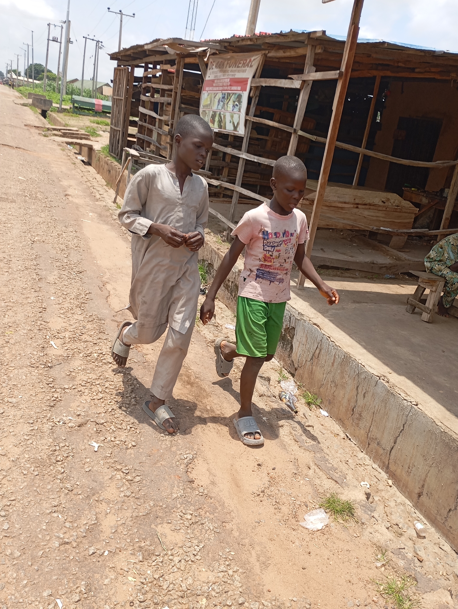Two young boys are walking down a dusty, unpaved road in Shendam, Nigeria. The boy on the left is wearing a loose-fitting, light-colored outfit, and the boy on the right is wearing a pink t-shirt and green shorts. They are walking side-by-side, with the boy on the left slightly ahead of the boy on the right. Behind them, on the right side of the road, is a makeshift structure made of wood and corrugated metal, possibly a market stall or workshop. The structure appears weathered and somewhat dilapidated. The ground is covered in dirt and small stones, with some patches of dry grass growing along the edge of the road. In the background, there are more structures, utility poles, and some trees, indicating a semi-urban or rural environment. The sky is overcast, suggesting a cloudy day. There is some debris, including plastic bottles and wrappers, scattered on the side of the road.