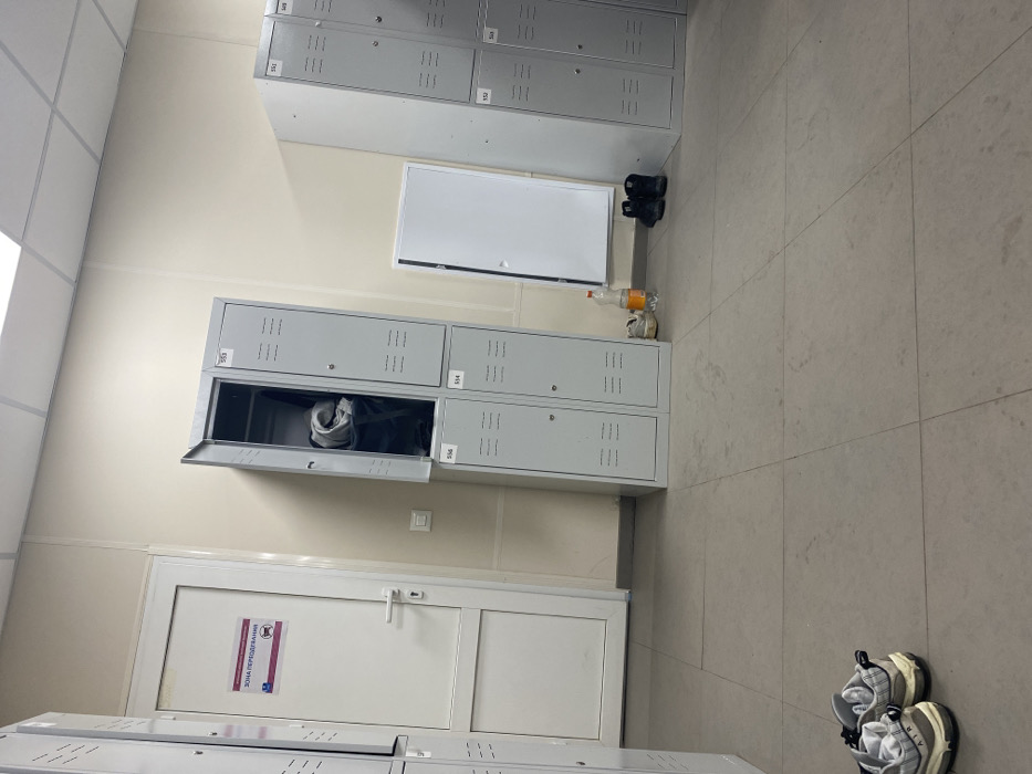 The image displays an interior view of a changing area located in Nikol'skoe, Russia. The room features light beige walls, a light grey tiled floor, and a white suspended ceiling with visible grid lines and integrated lighting.
On the left, a white interior door with a silver lever handle is visible. A rectangular sign with Russian text "ЗОНА ПЕРЕОДЕВАНИЯ" (Changing Area) and two graphic symbols is affixed to the door. Directly adjacent to the door, a stack of grey metal lockers is present, comprising four individual units arranged in two columns and two rows. The upper-left locker, numbered 555, is open, revealing dark fabric or clothing inside. The remaining three lockers in this stack are closed and numbered 556 (bottom left), 553 (top right), and 554 (bottom right). Each locker door features horizontal ventilation slats.
To the right of the central lockers, a white rectangular access panel or small door is partially ajar on the wall. Further to the right, another stack of grey metal lockers is partially visible, with doors numbered 551, 552, 550, and 549, all appearing closed.
On the floor, in the foreground to the left, a pair of light-colored athletic shoes with thick soles are positioned. Near the central locker unit, an orange plastic bottle stands next to a pair of light-colored casual shoes. Further right, a pair of black high-top shoes or boots are placed on the floor. No individuals are present in the scene.