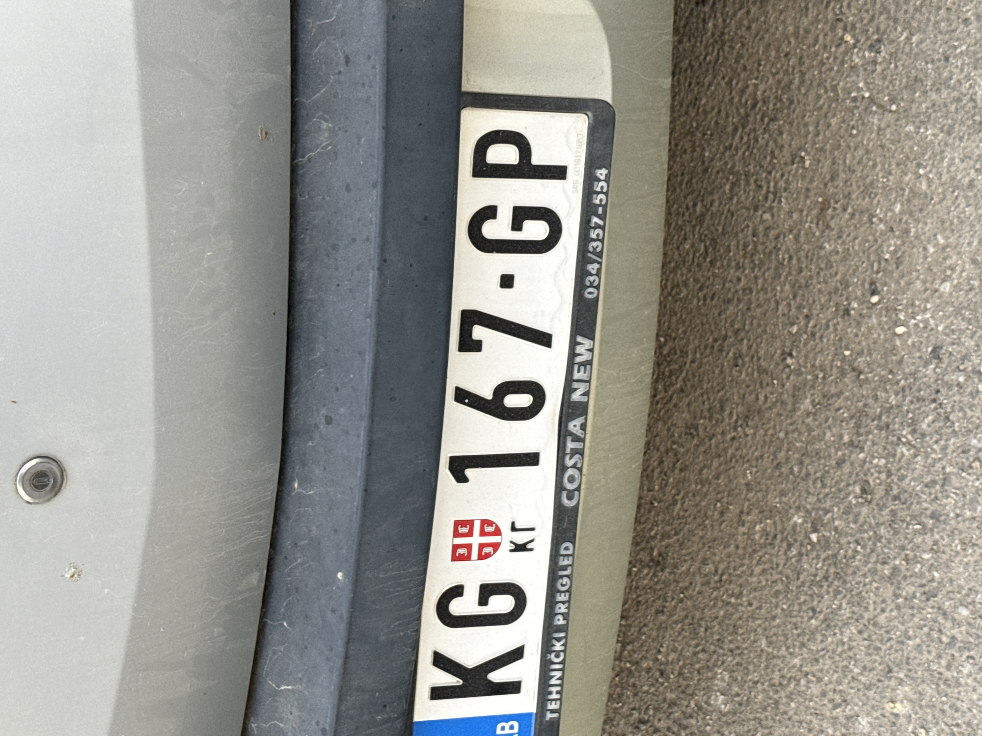 The image shows a Serbian license plate reading KG 167-GP. "KG" indicates the license plate is from Kragujevac, Serbia. The license plate frame contains the words "TEHNICKI PREGLED," the name "COSTA NEW," and a phone number, 034/357-554. The plate is attached to a light-colored vehicle. The background consists of a surface that looks like asphalt or concrete.