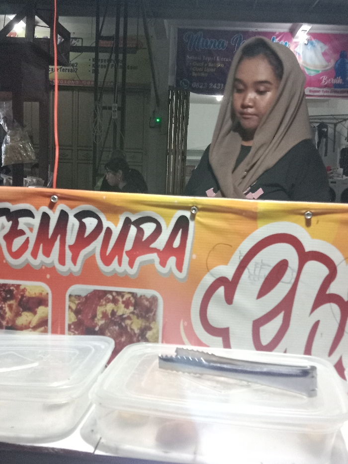 This image captures a nighttime scene at a food stall in Turunsih, Indonesia. A woman with medium-dark skin, wearing a dark long-sleeved shirt and a light brown hijab, stands behind a counter, positioned right of center and looking downwards. Partially visible in the left background is another individual, wearing a dark top, situated further back and appearing to look down or engage with something at a lower level.

In the foreground, a prominent yellow and orange banner with white trim spans the width of the image. Large red and white text on the banner reads "TEMPURA" on the left side, with two rectangular images of fried food items (possibly tempura or similar snacks) displayed below it. The right side of the banner shows a large, partially obscured red and white graphic.

On the white countertop below the banner, two clear rectangular plastic containers with lids are present. The container on the left appears mostly empty. The container on the right has a pair of silver metal tongs resting horizontally on its lid, and small, round, light brown food items are visible inside.

The background reveals elements of a commercial setting. To the left, an orange electrical cord hangs vertically. Behind the subjects, a light-colored double-door structure is visible. Various signs are mounted on the wall; one blue sign behind the woman advertises "Muna" with services like "Solusi Tepat Untuk..." and a phone number "0823 2431...". Another partial sign to the right shows "Berih" with laundry-related graphics. A green indicator light is visible on a wall or structure in the upper-center background, amidst various wires and structural components. The lighting suggests artificial illumination in a dark environment.