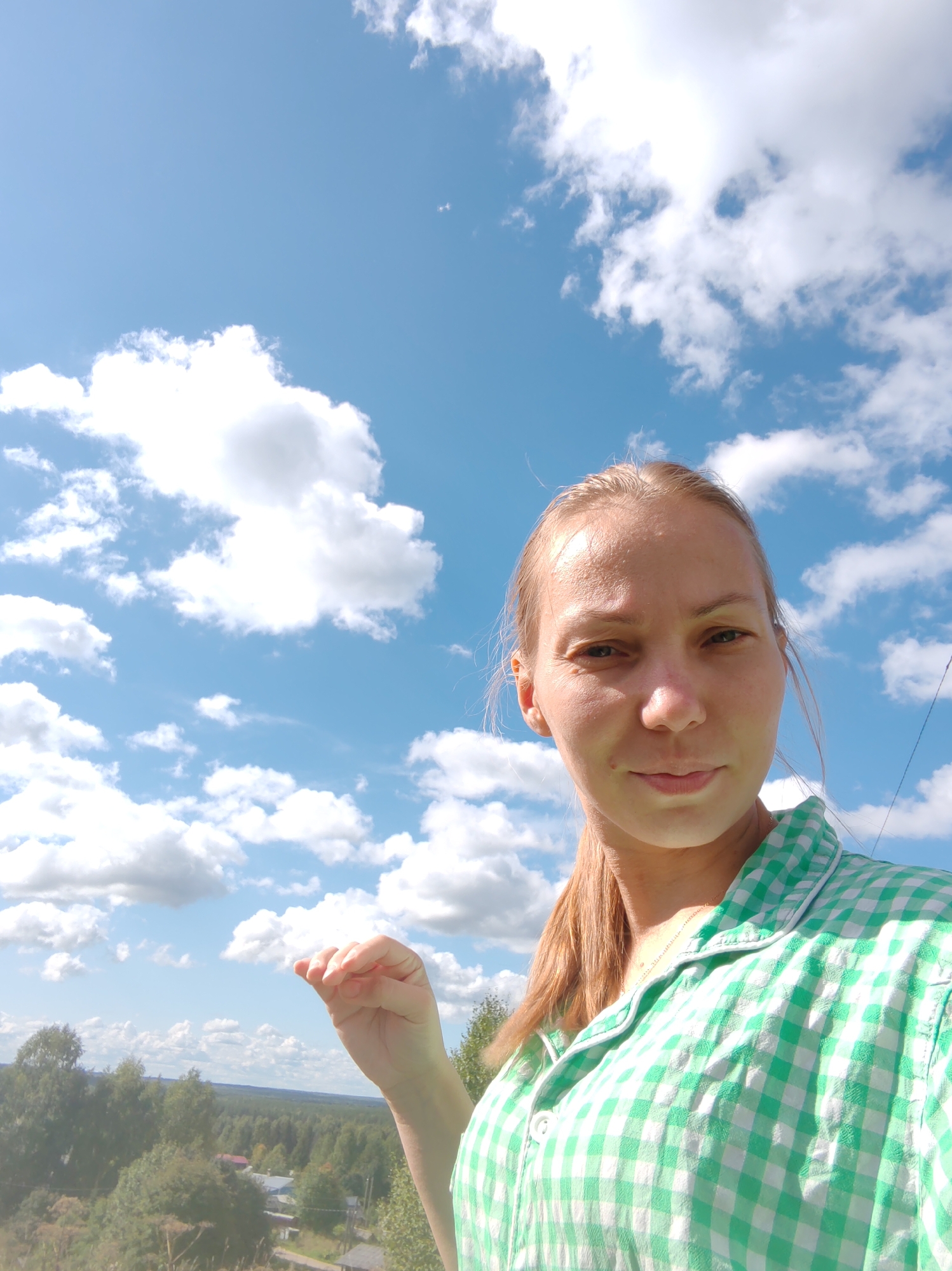 A young woman with blonde hair tied in a ponytail takes a selfie outdoors against a bright blue sky dotted with white clouds. She is wearing a green and white checkered shirt. Her right hand is raised, with fingers slightly curled, in the foreground. In the background, a dense forest stretches across a hilly landscape, with scattered houses visible amongst the trees, suggesting a rural or semi-rural location possibly near Чухлэм, Russia. The lighting indicates it is daytime, likely sunny.