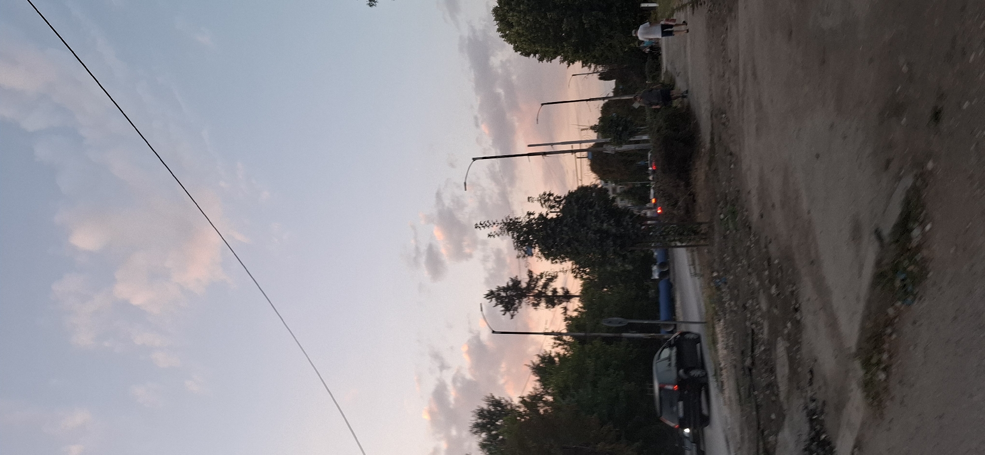 This image depicts a street scene in Pleven, Bulgaria, during what appears to be either dawn or dusk, given the soft, pastel hues of the sky and clouds. A thin, dark wire stretches diagonally across the upper portion of the frame. The foreground is a dirt and gravel path, littered with debris and patches of sparse vegetation. To the left, a paved road runs parallel to the path. On the road, a dark-colored station wagon is moving away from the camera. Beyond the road, a dense line of trees forms a backdrop, interspersed with streetlights and what appear to be utility poles. In the middle ground, a few individuals are visible walking along the paved area adjacent to the road. One person, wearing a white shirt and dark shorts, is walking towards the right, while another, in a dark shirt and shorts, walks in the same direction. The overall impression is of a quiet urban environment at the fringes of the day.