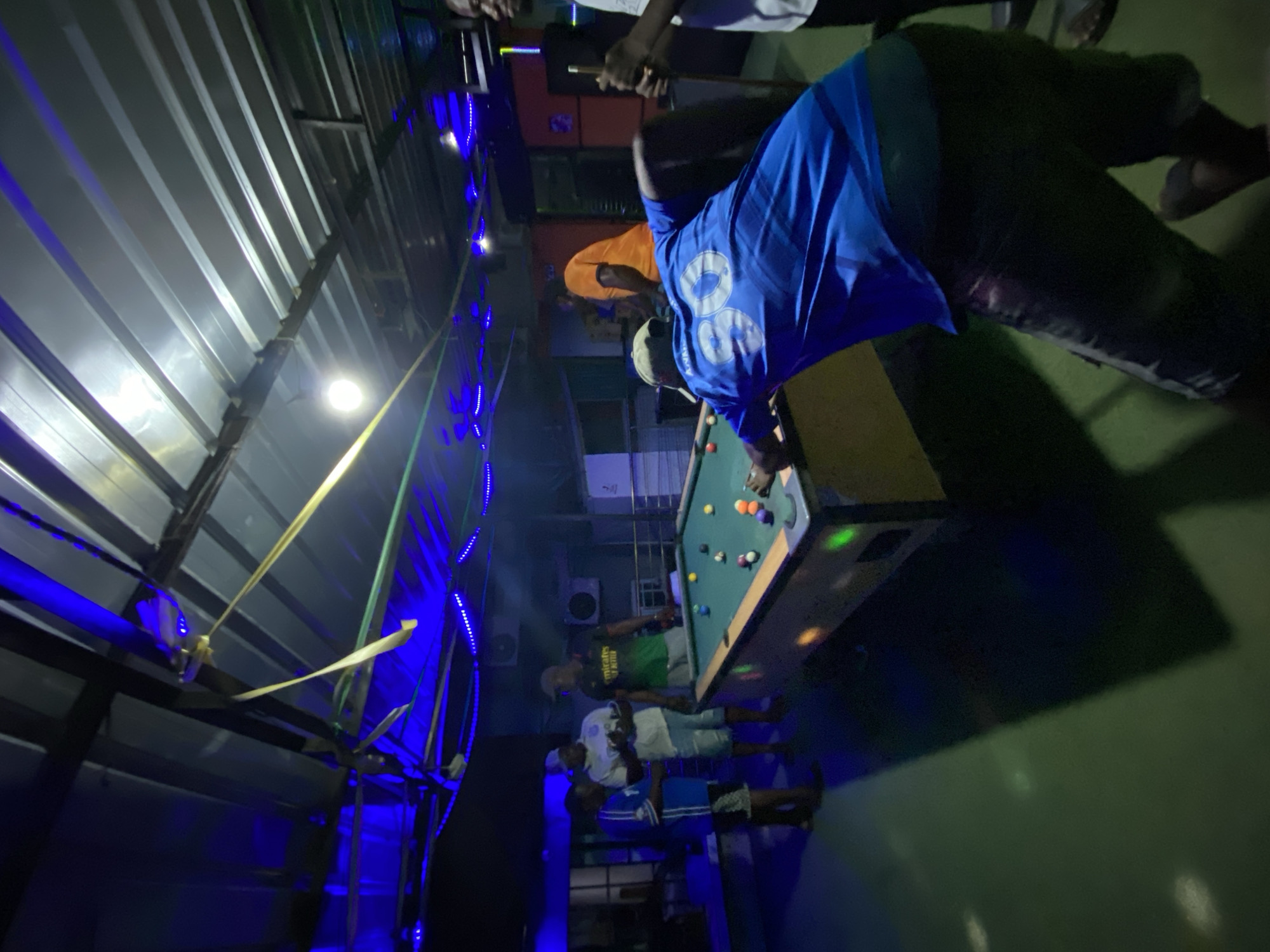 The image shows a group of people playing pool indoors in Ado, Nigeria. The building has a corrugated metal ceiling decorated with blue LED strip lights. One man in a blue jersey is leaning over the pool table, lining up a shot. Other people are standing around watching the game. Air conditioning units are mounted on a wall in the background.