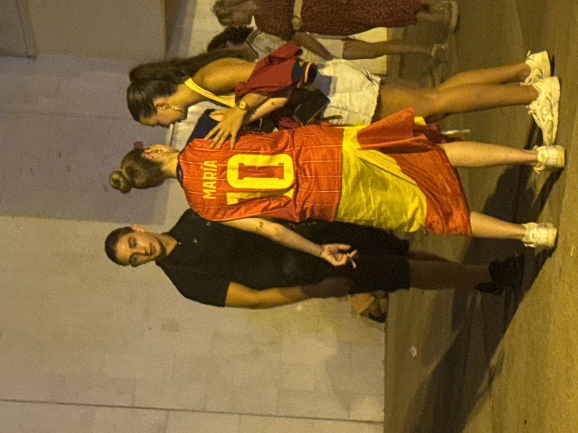 In Blanes, Spain, at night, three individuals are standing near a wall, with two women in the foreground. The woman on the left wears a red and yellow Spanish national soccer team jersey with "MARIA" and the number "10" printed on it, and a Spanish flag draped around her lower body. The woman next to her, on the right, wears a yellow tank top and white shorts. To their left stands a man in a black polo shirt, holding a cigarette in his right hand. The background is a textured, illuminated wall, and the ground appears to be paved. In the background, other people are visible, including a woman in a red patterned dress.