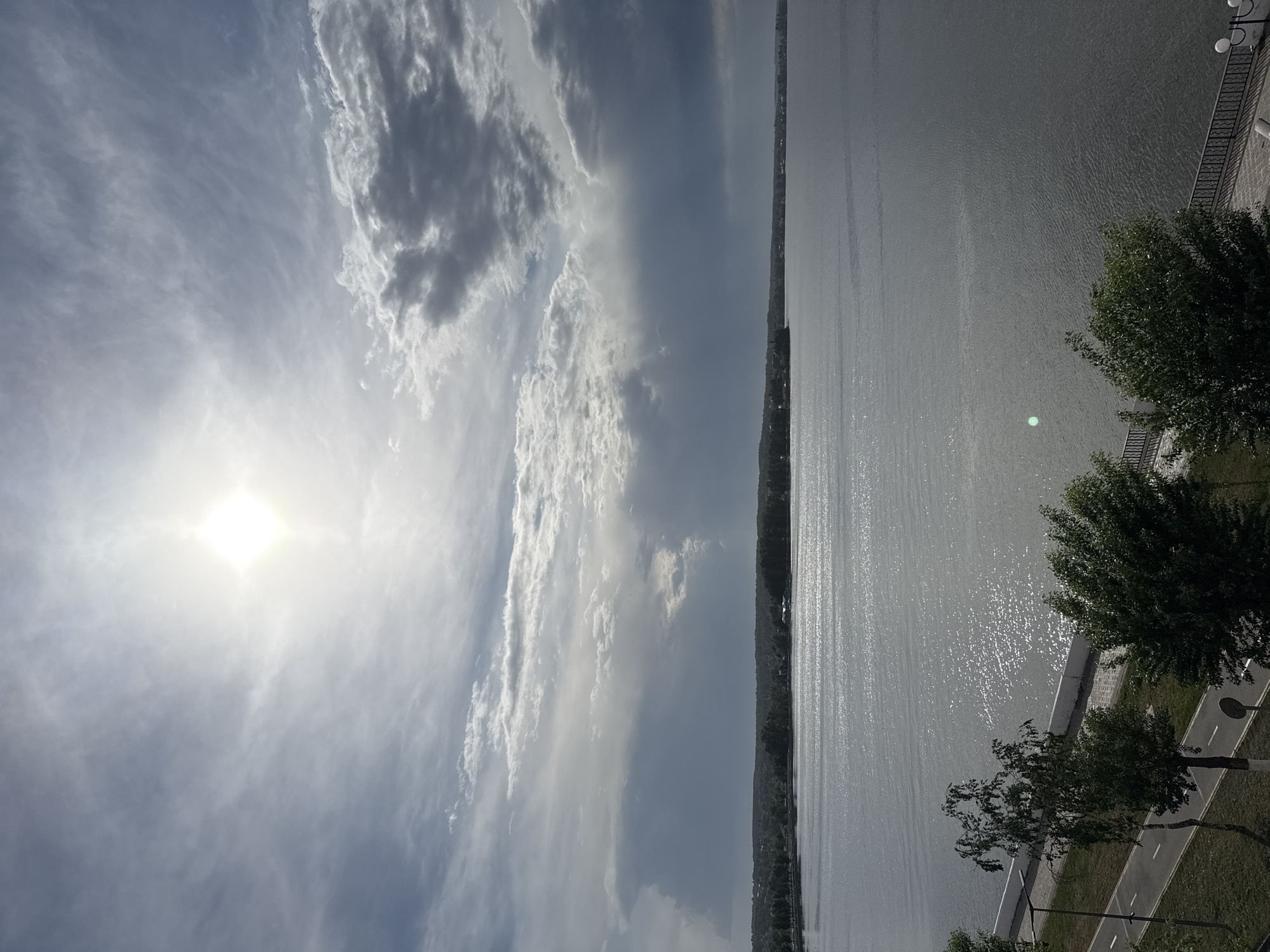 The image shows a body of water, likely the Votkinsk Reservoir, bordering a tree-lined embankment and a walkway in Votkinsk, Russia. The sky is visible, with scattered clouds and the sun prominently shining through. The water surface reflects the sunlight, creating a shimmering effect. Trees are visible along the edge of the water, next to the embankment and walkway. A lamppost is located near the path. The perspective is an elevated, angled view.