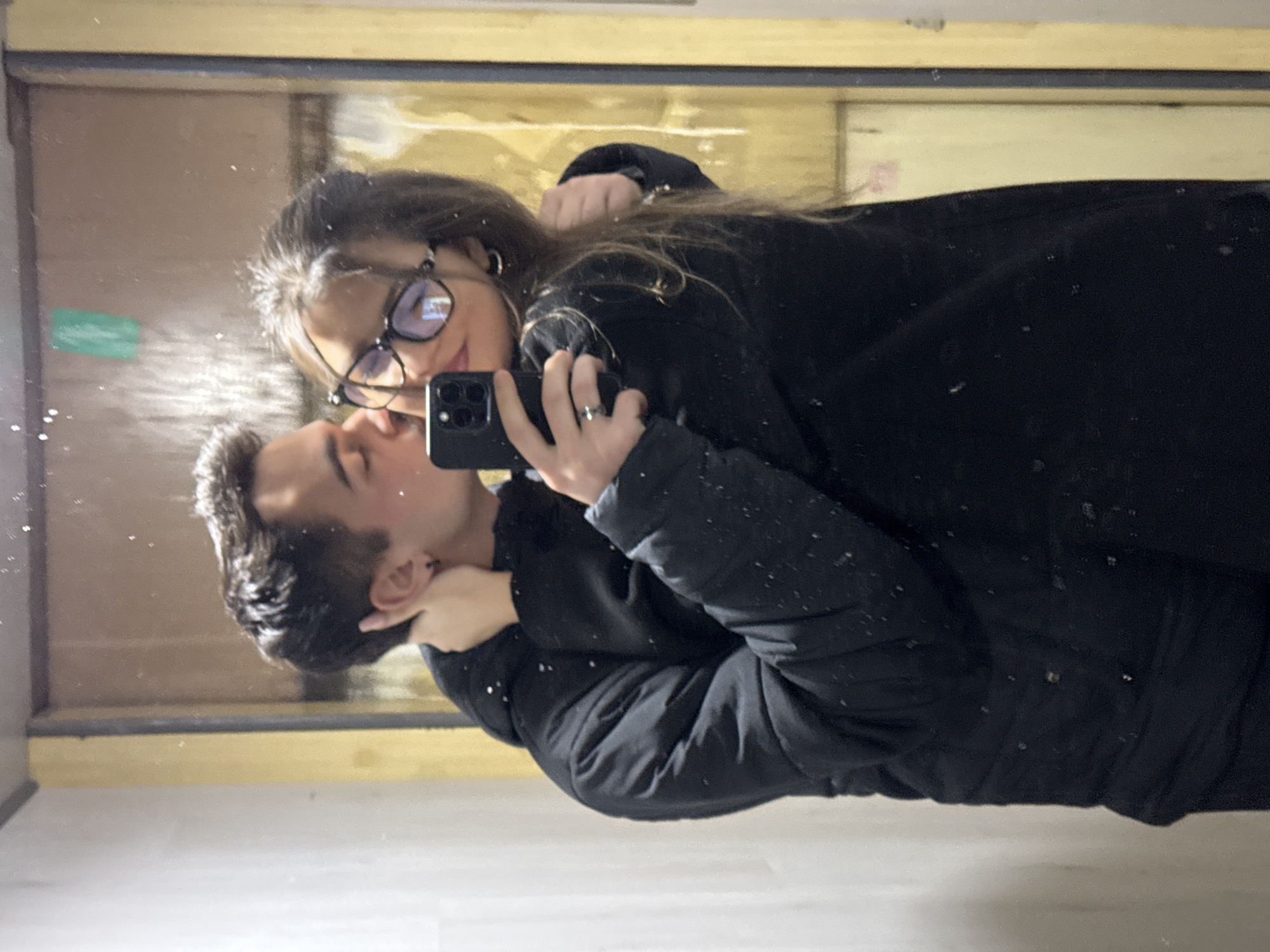 A mirror selfie captures two individuals, a male and a female, engaged in an affectionate interaction within an indoor setting. The male, positioned on the left side of the reflection, wears a black puffer jacket and has short, dark hair. He is shown kissing the female on her right cheek, with his left arm around her neck. A small, dark earring is visible in his left ear. The female, on the right, wears black-rimmed glasses and a dark jacket, with her long, dark hair styled down. She holds a black smartphone, consistent with an iPhone model, in her right hand to take the photograph. A ring is visible on the fourth finger of her right hand, and her left arm is around the male's waist. The mirror surface exhibits numerous small dust particles or smudges. The reflected background consists of a light, off-white wall on the left and a darker, yellowish-brown panel or door frame on the right. A small green rectangular sticker is partially visible on the upper right side of the reflected background. The specific location within Sofia, Bulgaria, cannot be determined from the visual information provided.