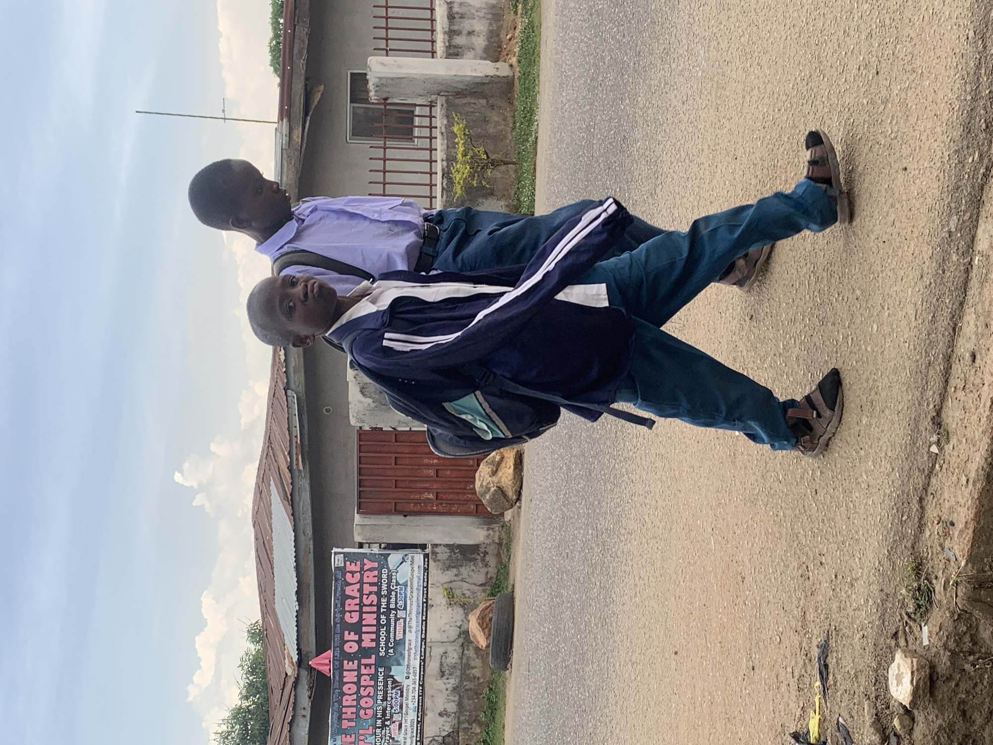 Two young boys walk along a paved road in Gura Top, Nigeria. The boy in the foreground wears a navy and white jacket and blue jeans with dark sandals. He has a backpack. The boy behind him wears a light purple shirt and dark blue pants. A sign for "Throne of Grace Gospel Ministry" is visible on the side of a building. The building has a corrugated metal roof.