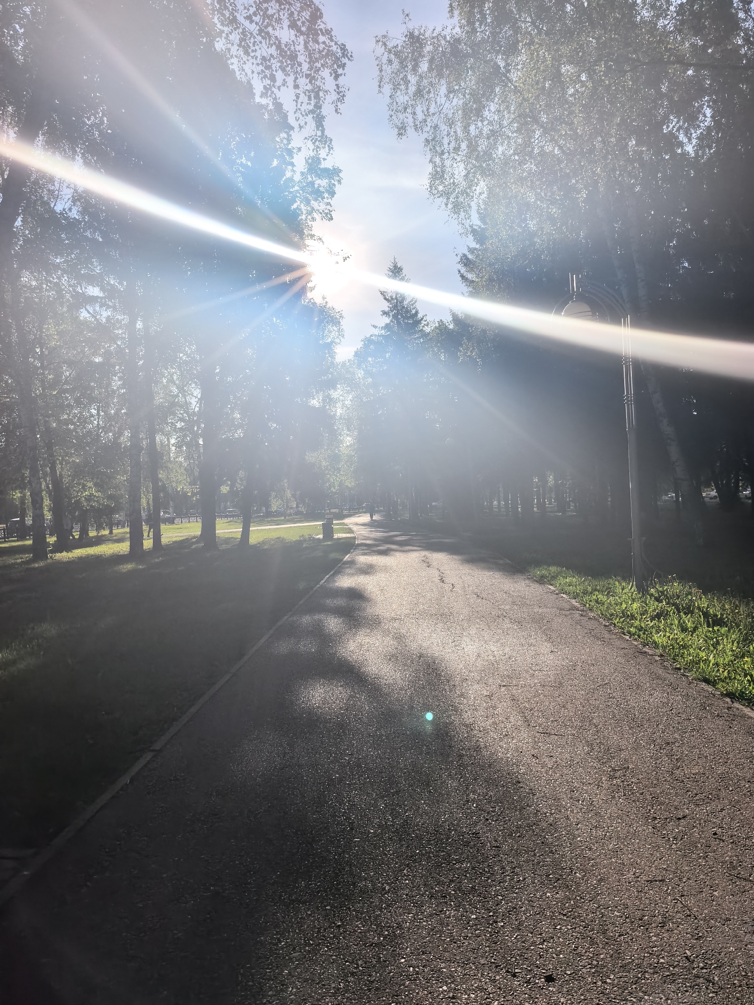 The image shows a pathway in a park in Sterlitamak, Russia. The path is paved with asphalt and stretches into the distance, curving slightly. Trees line both sides of the path, and sunlight streams through their leaves, creating bright flares in the image. A street lamp is visible on the right side of the path. A person is walking in the distance on the path. The grass is green along the edges of the path.