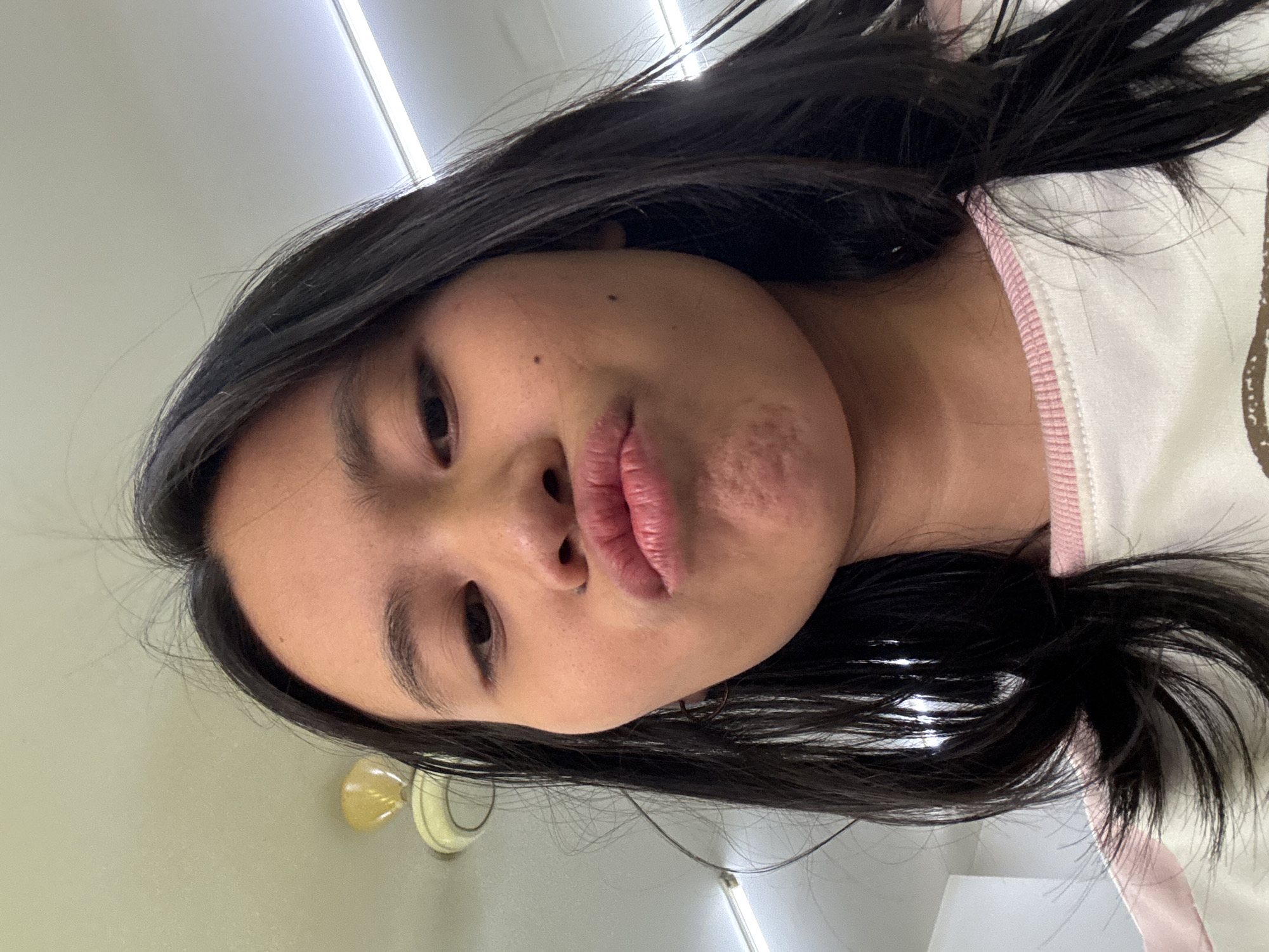 A young Asian woman with long, dark hair is captured in a close-up selfie, puckering her lips. She is wearing a white and pink collared shirt. The background features a white ceiling with fluorescent lighting fixtures and a yellow ceiling fan. The scene appears to be indoors, likely in a residential or common area. The overall lighting suggests daytime.