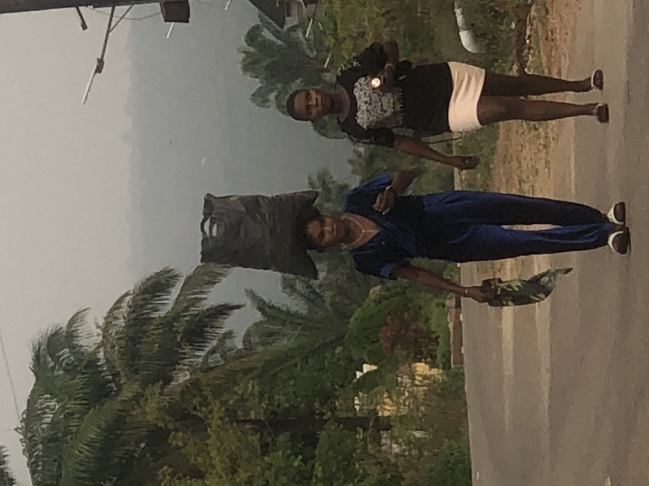 The image depicts two adult females walking on a paved road in Uke, Nigeria, under overcast or hazy daylight conditions. The woman positioned in the foreground, left of center, wears a dark blue jumpsuit or coordinated top and wide-legged pants, along with light-colored open-toed sandals. She carries a large, dark gray or black bag balanced on her head and a folded piece of patterned fabric in her right hand. A gold-toned necklace and wrist adornment are visible. Her gaze is directed slightly forward-left. The second woman, walking to the right and slightly behind the first, wears a black t-shirt with a white graphic design on the front, a light peach-colored short skirt, and dark open-toed sandals. She holds a small, light-colored cylindrical object, possibly a flashlight, in her right hand, and her gaze is directed forward. The road surface is asphalt and appears to curve gently to the left. Dense tropical vegetation, including numerous palm trees and other green foliage, lines both sides of the road. A hazy, grayish sky dominates the background, obscuring distant hills or mountains. A utility pole with multiple cross arms and wires is visible on the upper right side of the frame, and a light-colored structure is partially visible behind the foliage on the far left. White cylindrical objects are present on the ground level near the right edge.