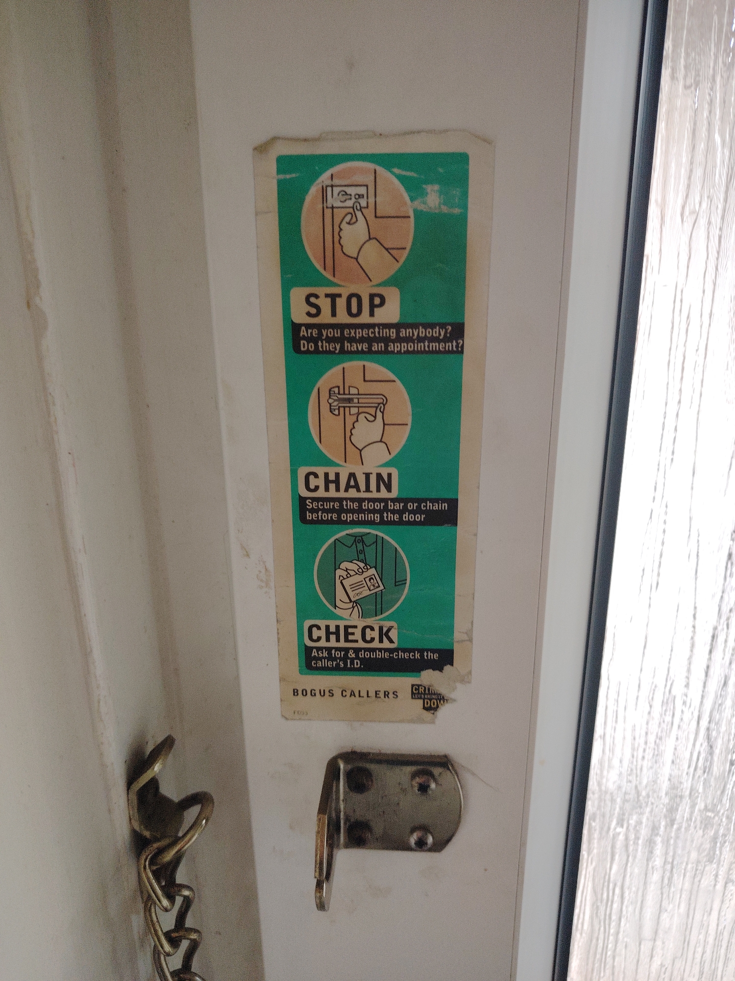 A vertical security poster, showing three illustrated steps for door safety, is affixed to a white door in Huntingdonshire, United Kingdom. The poster, displaying signs of age and wear with peeling edges and fading colors, is predominantly teal green with orange and white accents.
The top section, labeled "STOP," features an illustration of a hand pressing a doorbell or intercom button, accompanied by the text: "Are you expecting anybody? Do they have an appointment?"
The middle section, labeled "CHAIN," shows a hand engaging a door chain or bar mechanism. The accompanying text reads: "Secure the door bar or chain before opening the door."
The bottom section, labeled "CHECK," depicts a hand holding an identification card up, with a partial view of a person's shirt behind it. The text advises: "Ask for & double-check the caller's I.D."
Below the main instructions, the poster displays "BOGUS CALLERS" in large letters, and partially visible smaller text below that reads "CRIM" and "DOWN" from "LET'S BRING IT DOWN," along with a code "F095."
To the lower left of the poster, a brass-colored metal door chain is visible, hanging from its fixed bracket. To the lower right of the poster, a silver-colored metal bracket, likely for a door bar or a similar security device, is mounted with two screws. The door itself appears to be white, and a section of textured, possibly privacy, glass is visible along the right edge of the frame.